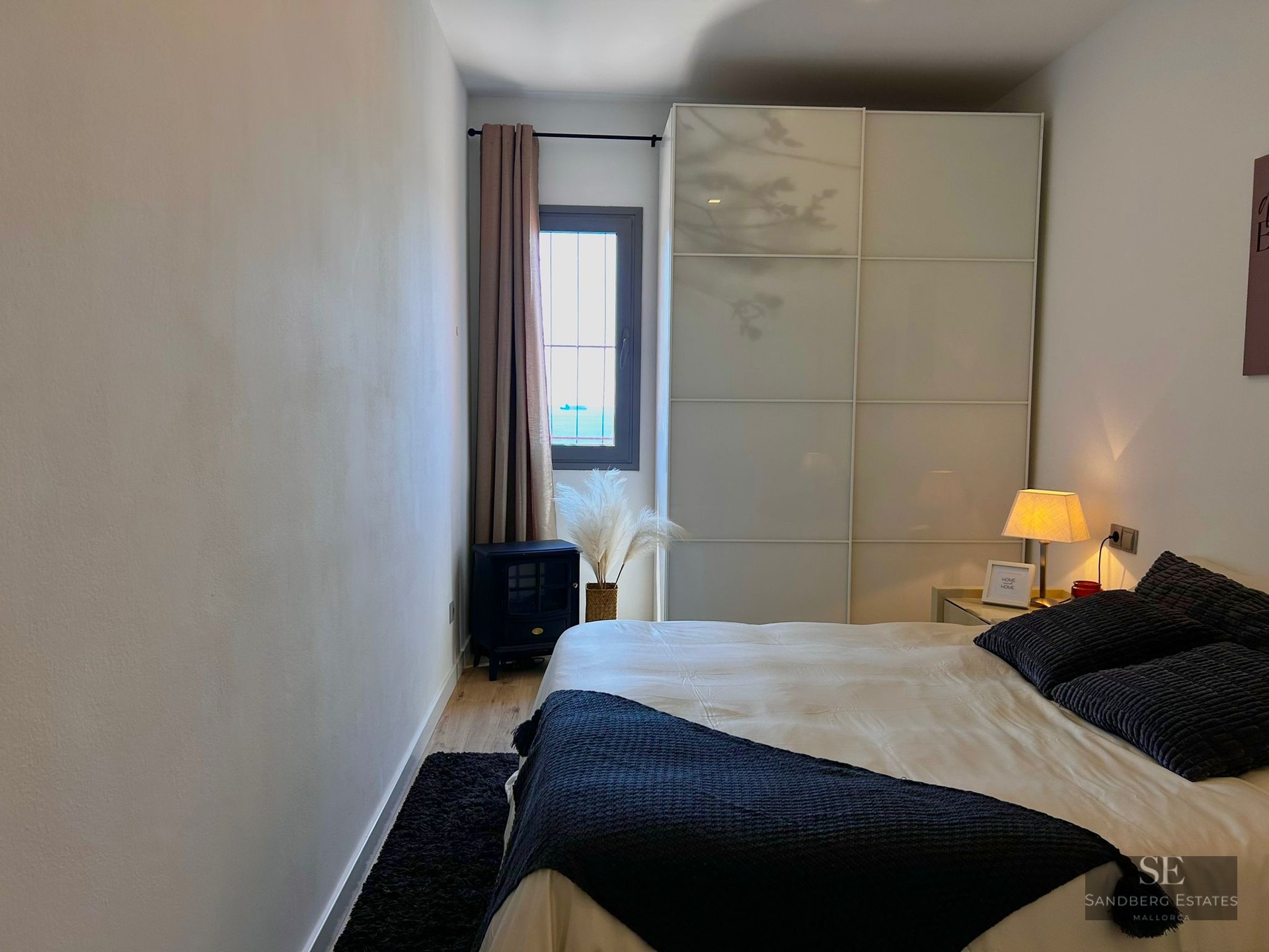 Modern bedroom featuring a large white wardrobe, a bed with dark pillows, and a small window with a sea view.