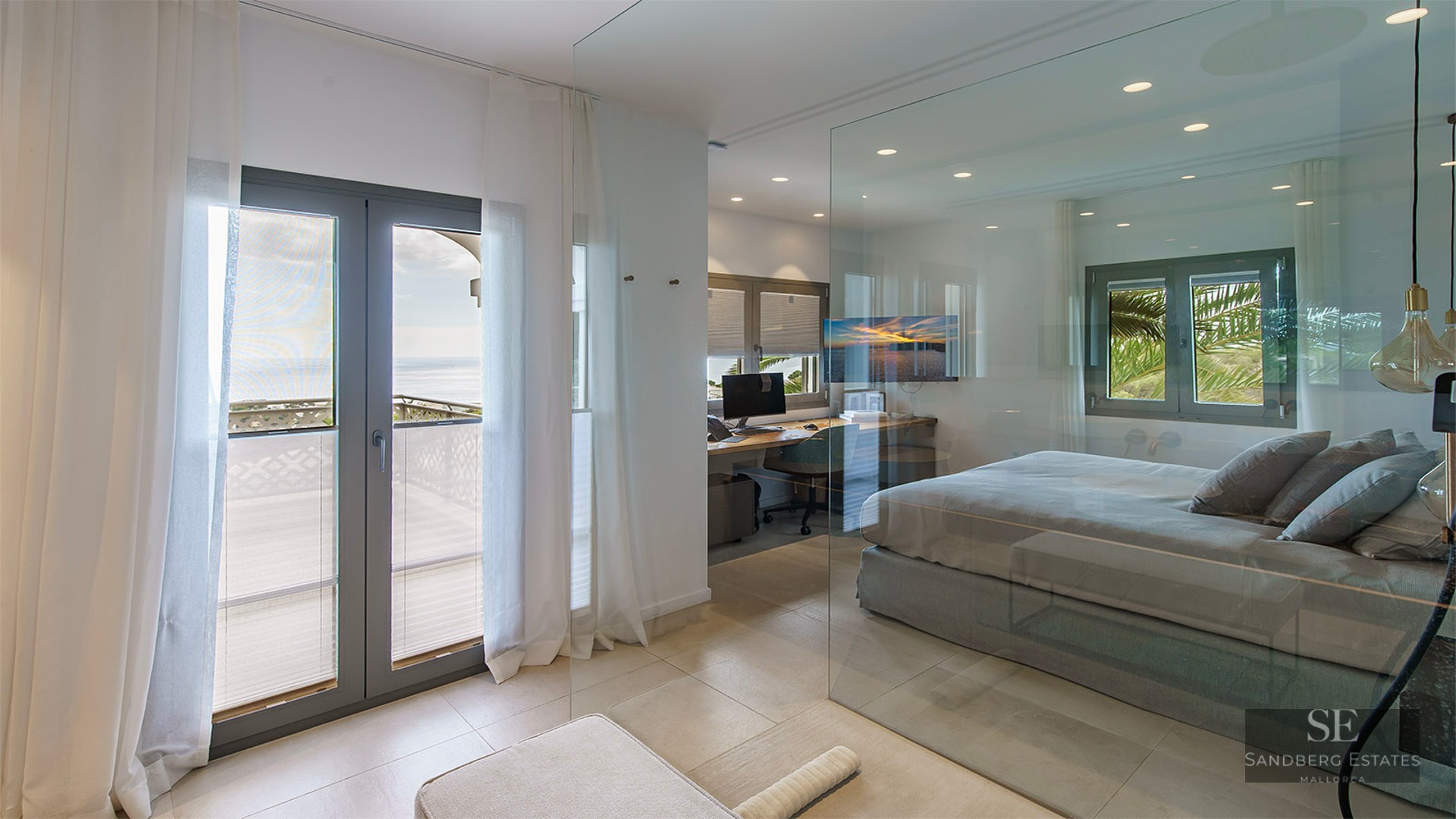 Modern bedroom with floor-to-ceiling glass wall, integrated office area, and balcony overlooking the sea.