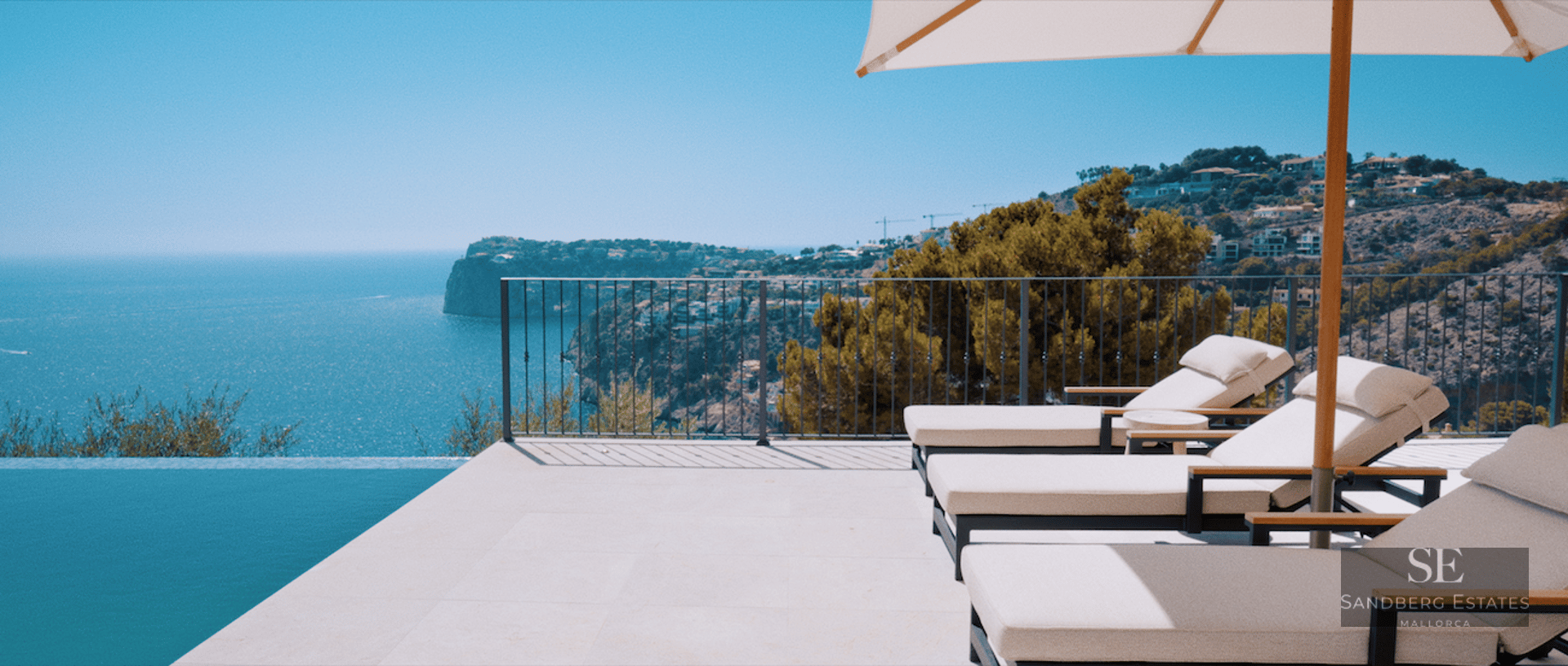 Luxury terrace featuring white sun loungers and a parasol next to an infinity pool overlooking the sea and cliffs.