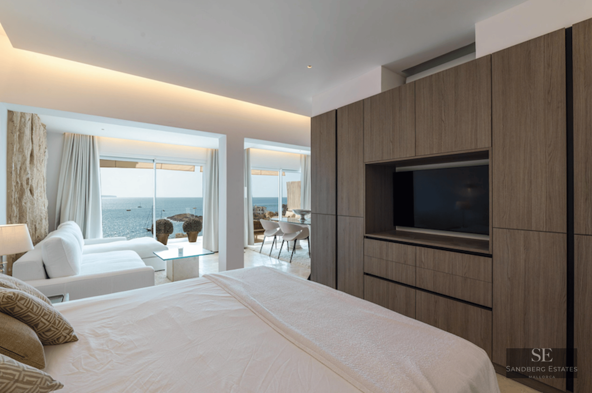 Master bedroom featuring a large wooden wardrobe with TV, white bed, and floor-to-ceiling windows overlooking the ocean.
