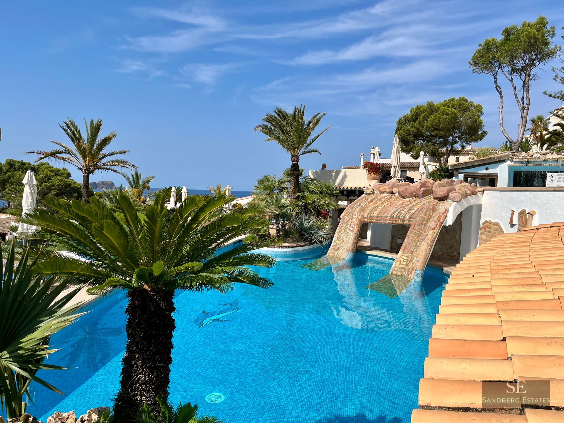 Large blue swimming pool surrounded by palm trees and Mediterranean architecture with a view of the sea in the distance.