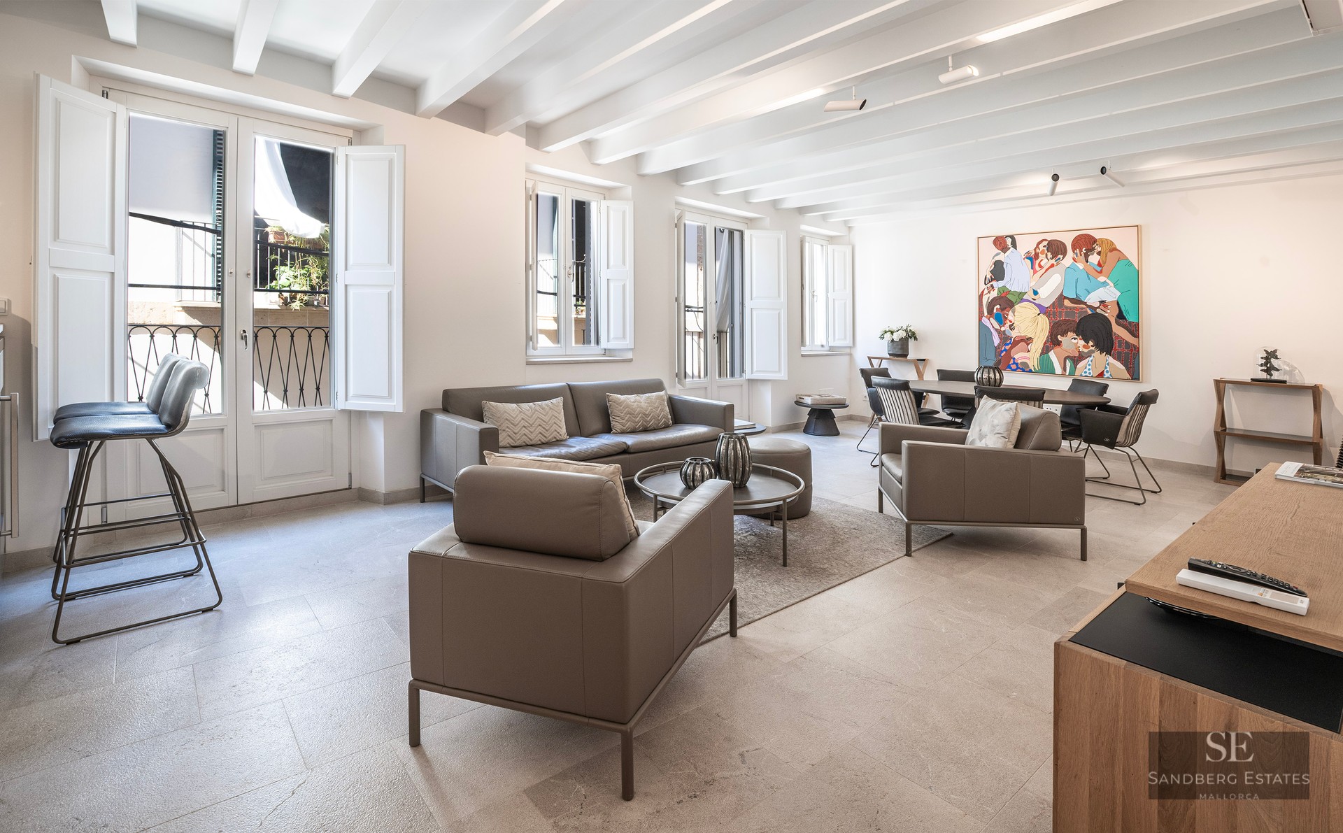 Salon lumineux avec canapés en cuir gris, plafond aux poutres blanches et coin repas avec une œuvre d'art colorée.