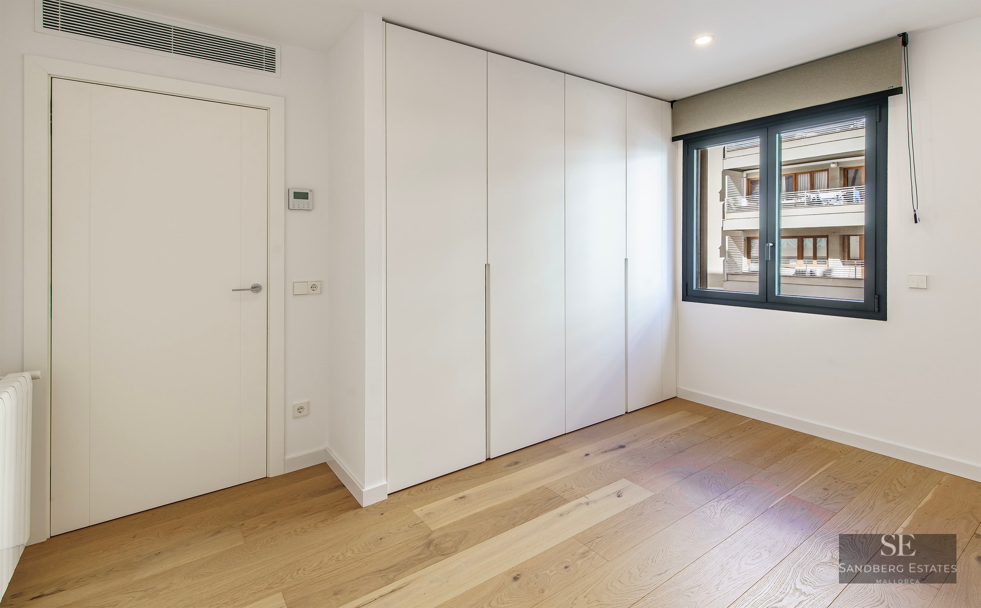 Chambre lumineuse avec parquet en bois clair, grands placards blancs intégrés et fenêtre au cadre sombre.