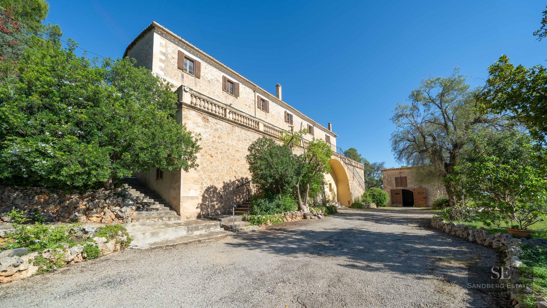 Grand stone Mediterranean villa with an elevated terrace, wooden shutters, and lush trees against a clear blue sky.