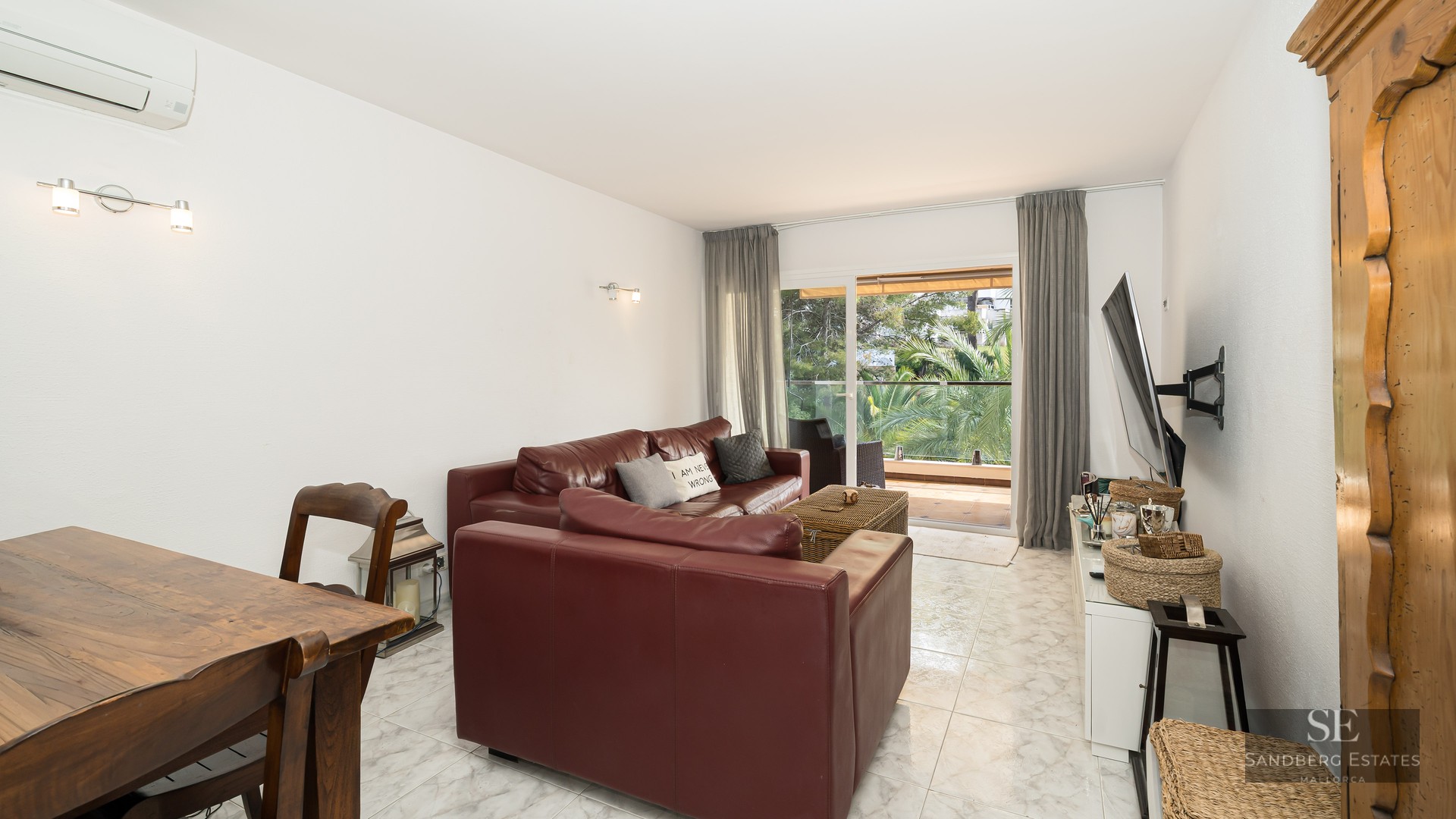Modern living room featuring burgundy leather sofas, white marble floors, and a glass door view to a lush green balcony.