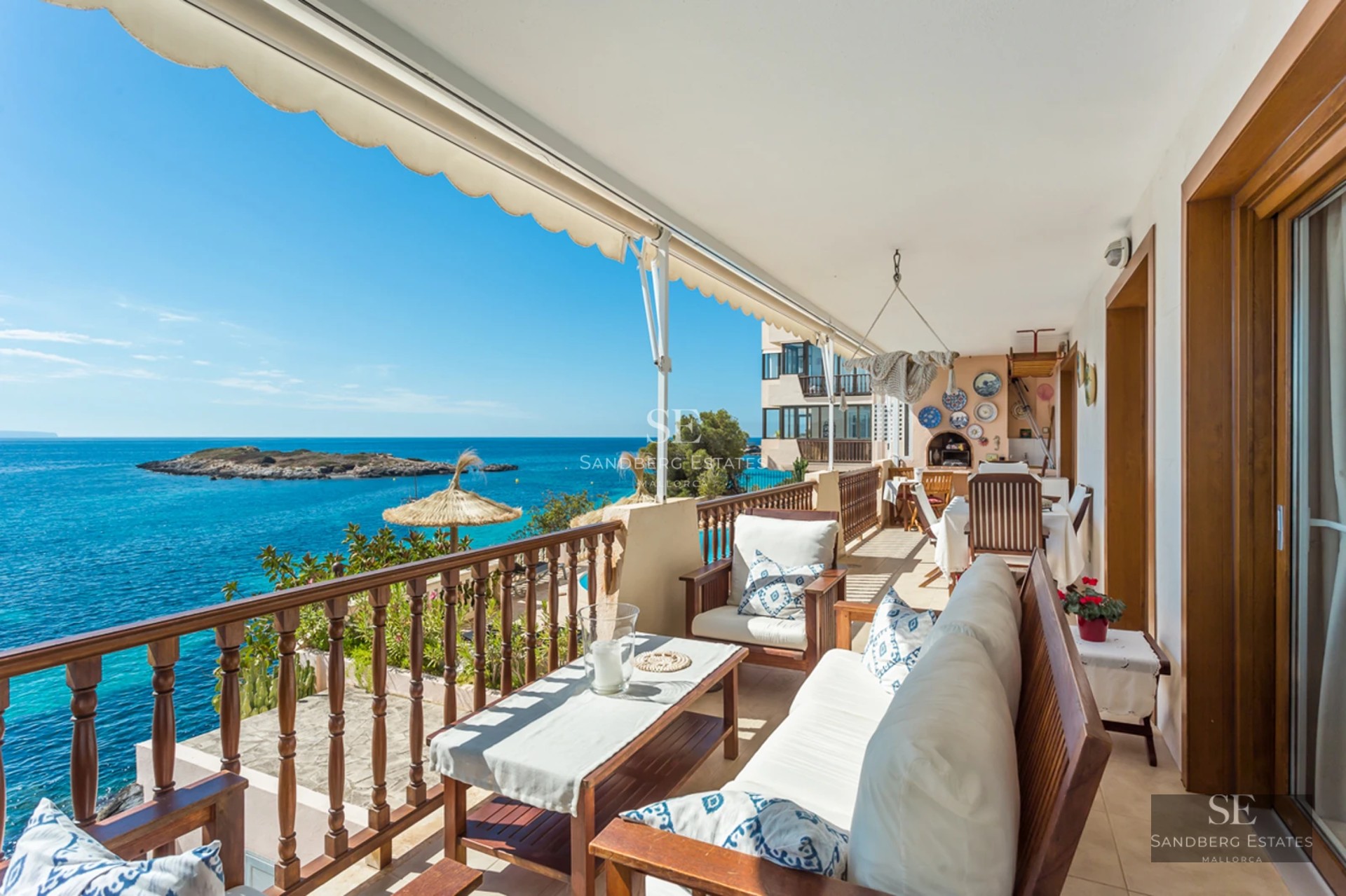 A wooden terrace with white sofas and a dining table overlooking a turquoise sea and a small island.