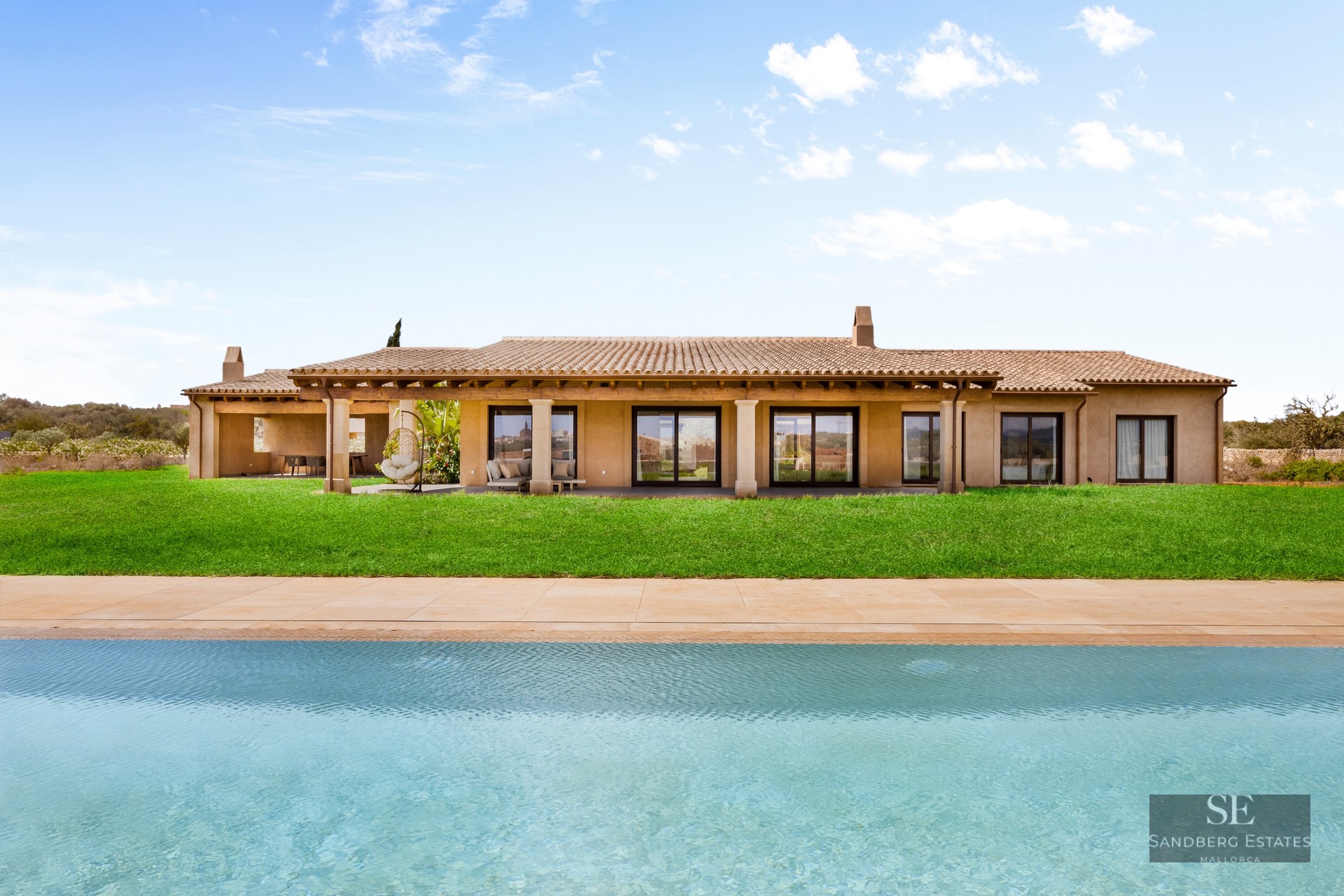 Large turquoise pool in front of a modern Mediterranean villa with a green lawn and terracotta roof under a blue sky.