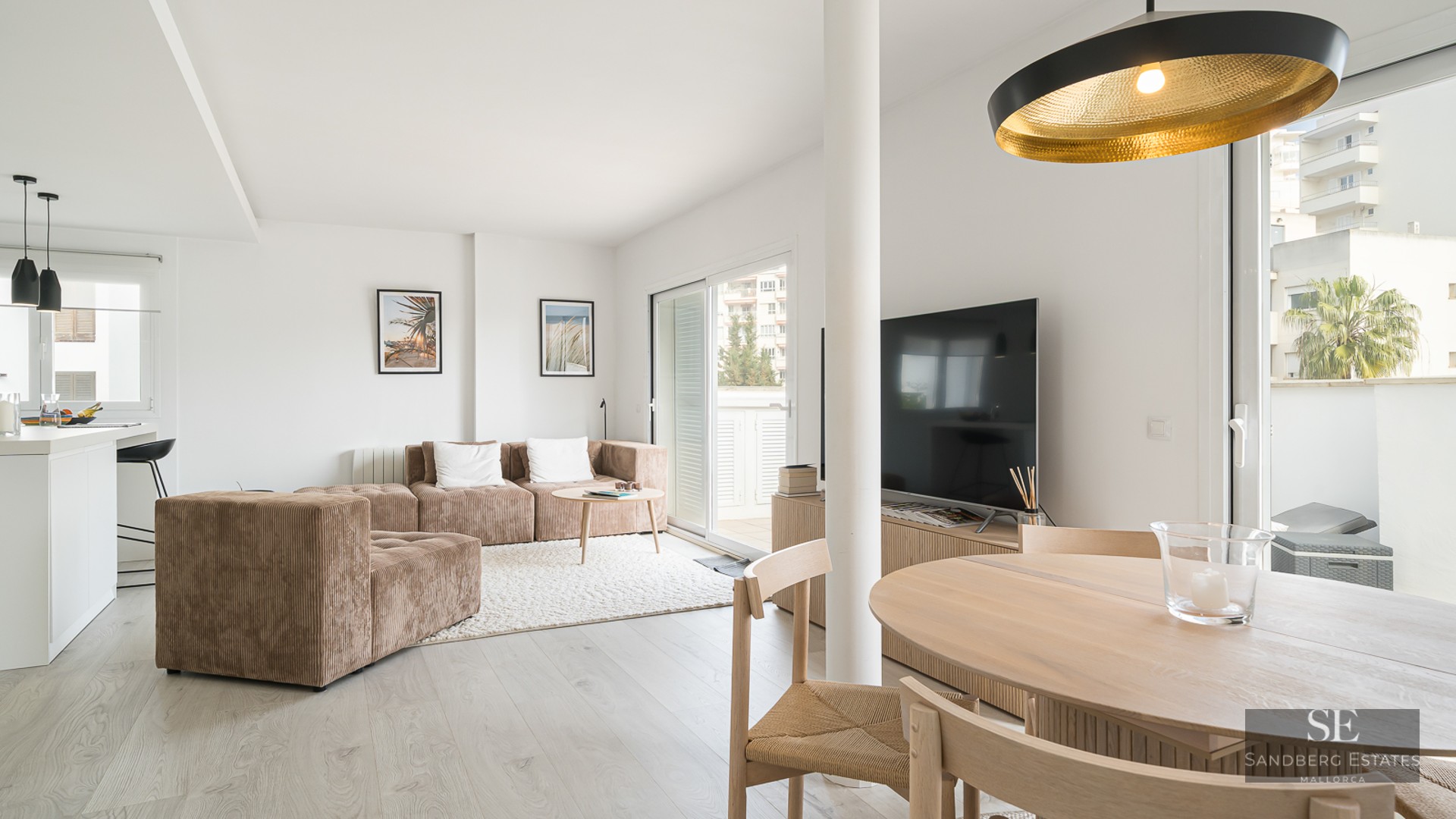 A sunlit living room featuring a brown modular sofa, wooden dining table, and large glass sliding doors leading to a balcony.