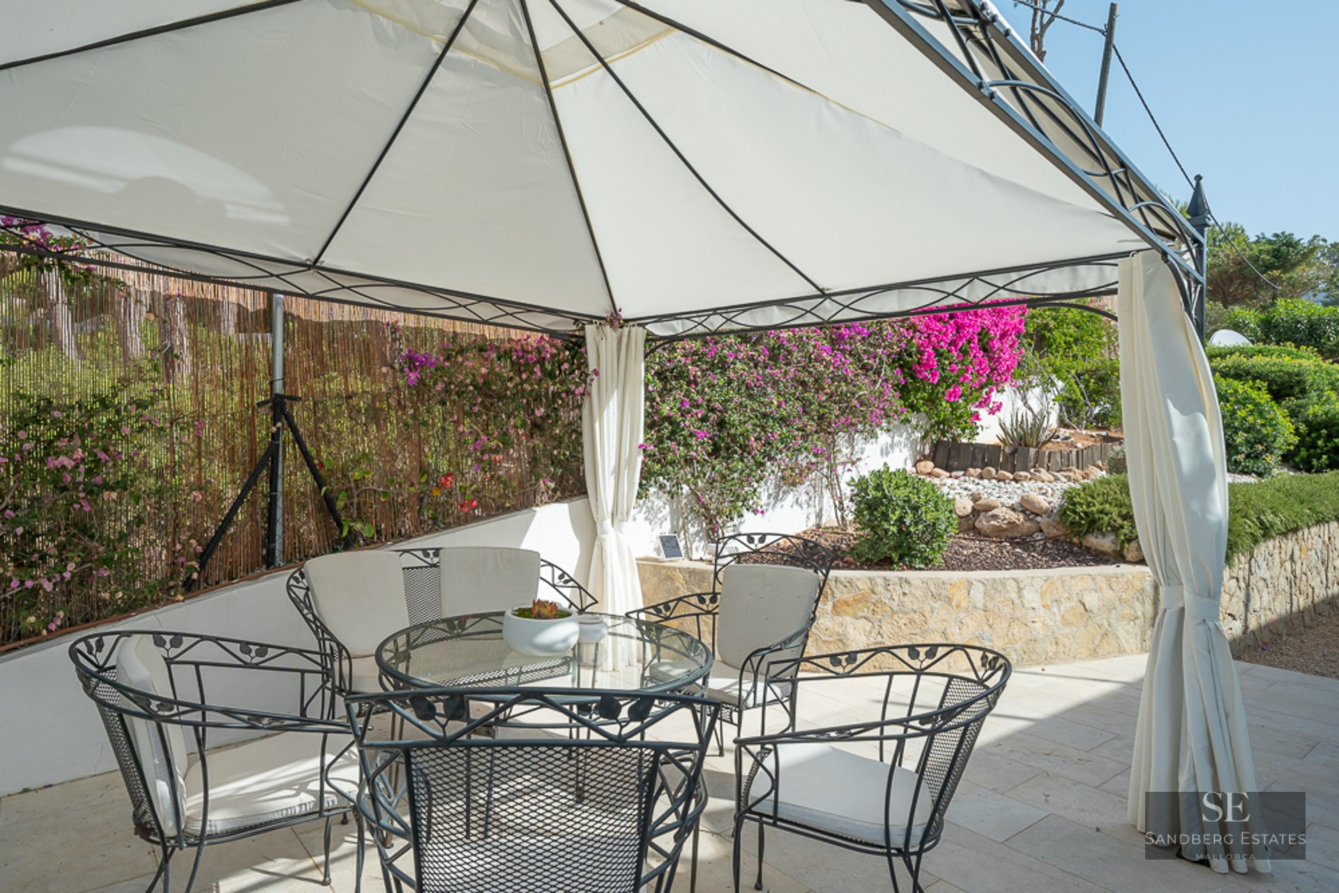 A white gazebo with a glass table and black wrought-iron chairs on a stone patio surrounded by pink flowers.