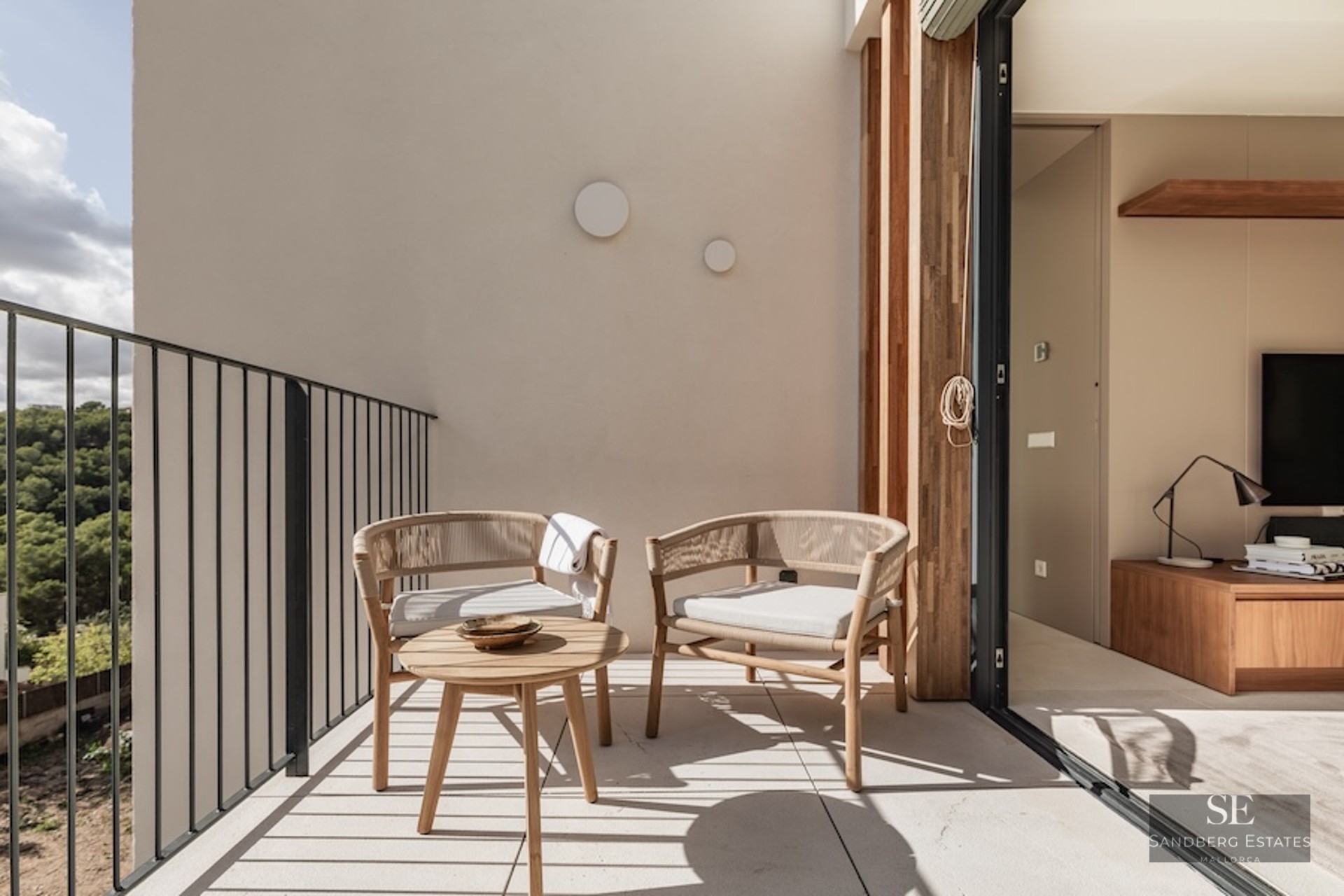 Sunlit balcony with designer woven chairs and a wooden coffee table overlooking a lush green forest.