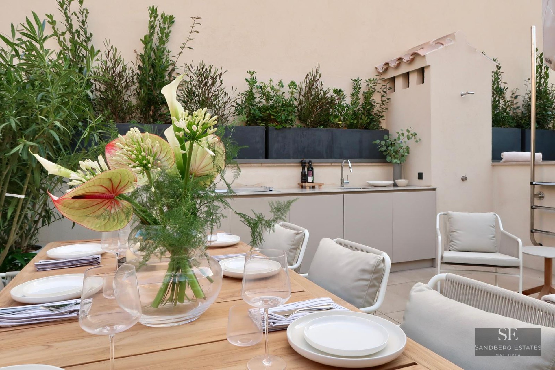 A wooden dining table set for a meal next to a modern outdoor kitchen and lush green planters.