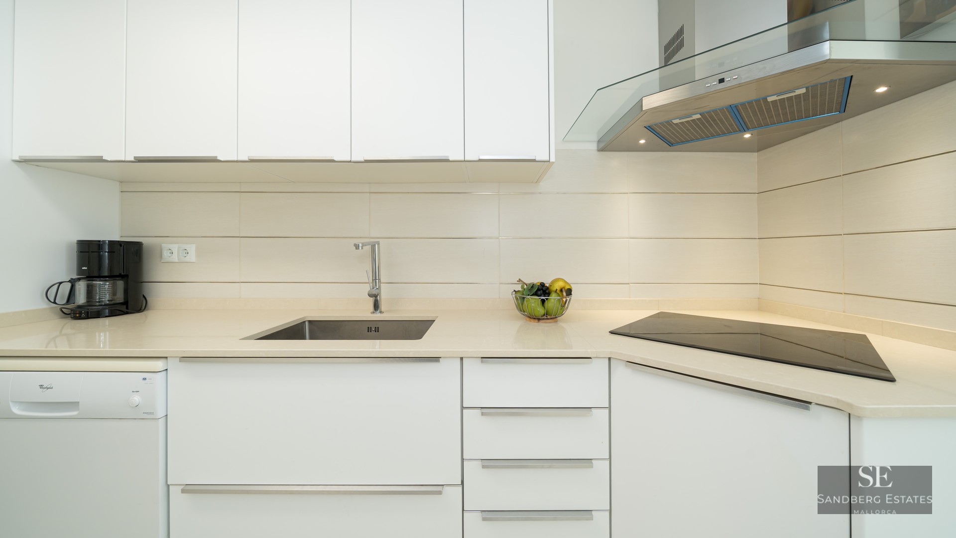 Close-up of a modern white kitchen featuring a stainless steel sink, glass hood, and ceramic cooktop.