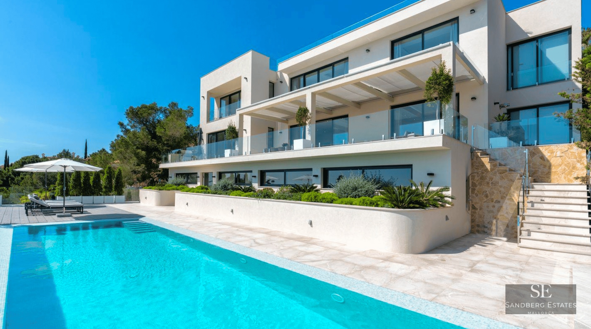 Large turquoise swimming pool in front of a modern white three-story villa with glass balconies and stone stairs.