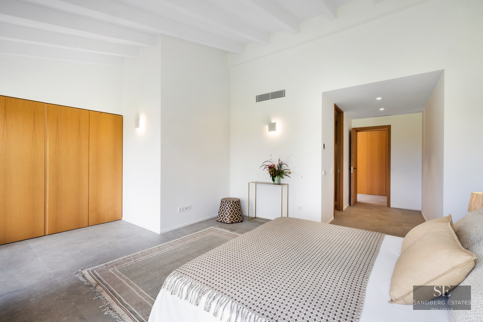 Bright bedroom with white beamed ceilings, wooden built-in wardrobes, large bed with beige pillows, and grey stone flooring.