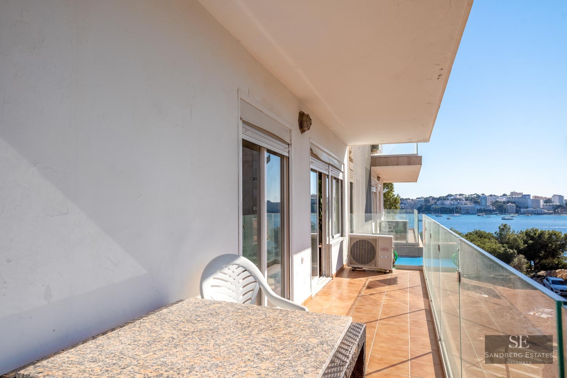 Balcony with glass railing and stone table overlooking a blue bay with boats and coastal buildings.
