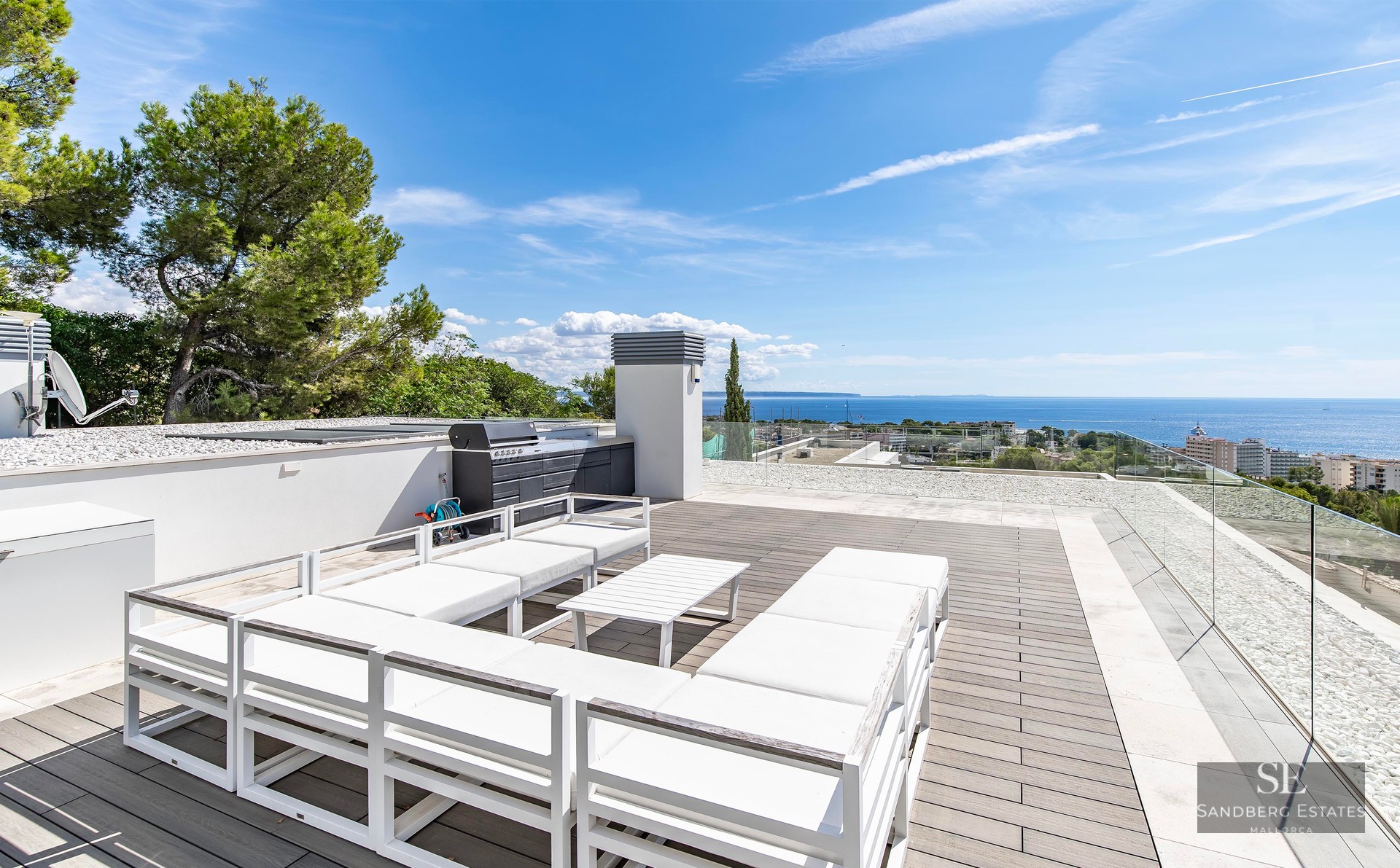 Spacious modern rooftop terrace with white lounge seating, glass railings, and a wide view of the sea under a blue sky.
