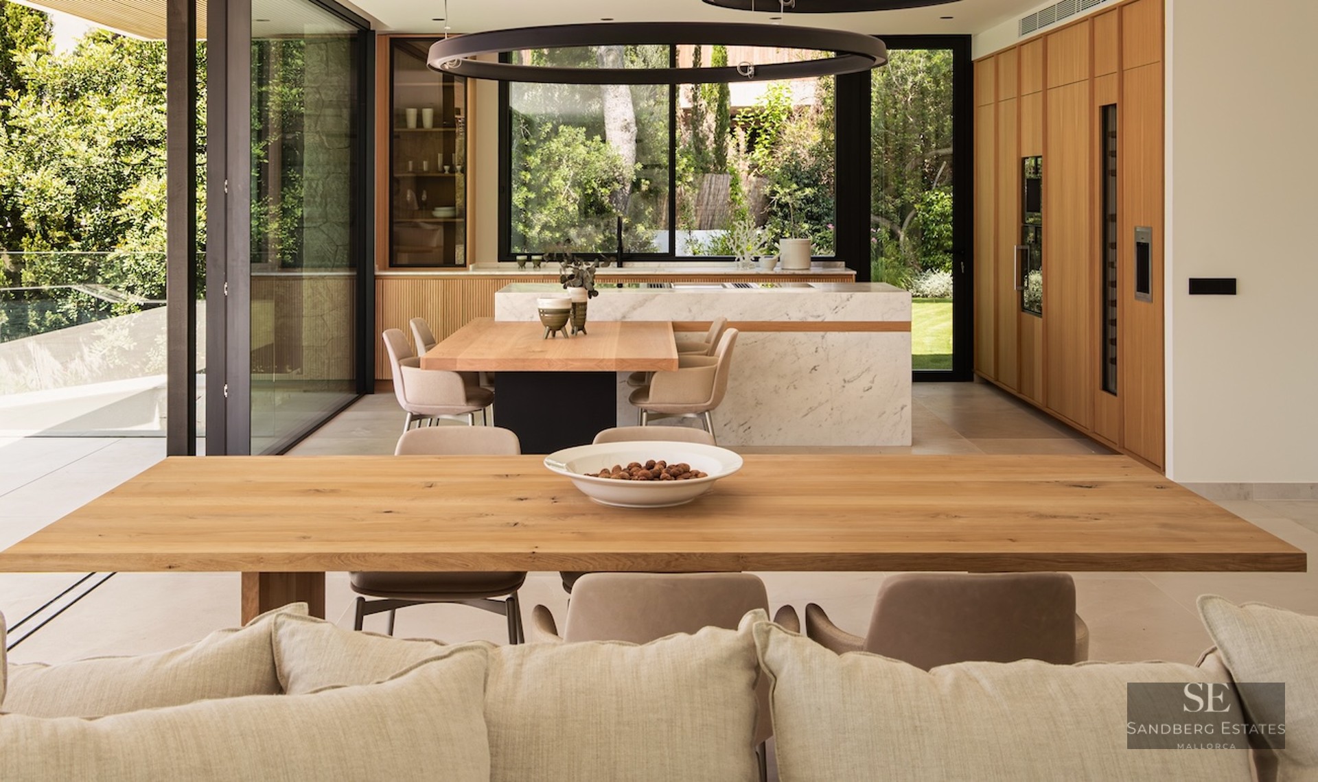 Modern kitchen with a large marble island, integrated wooden dining tables, and floor-to-ceiling windows showing a garden.