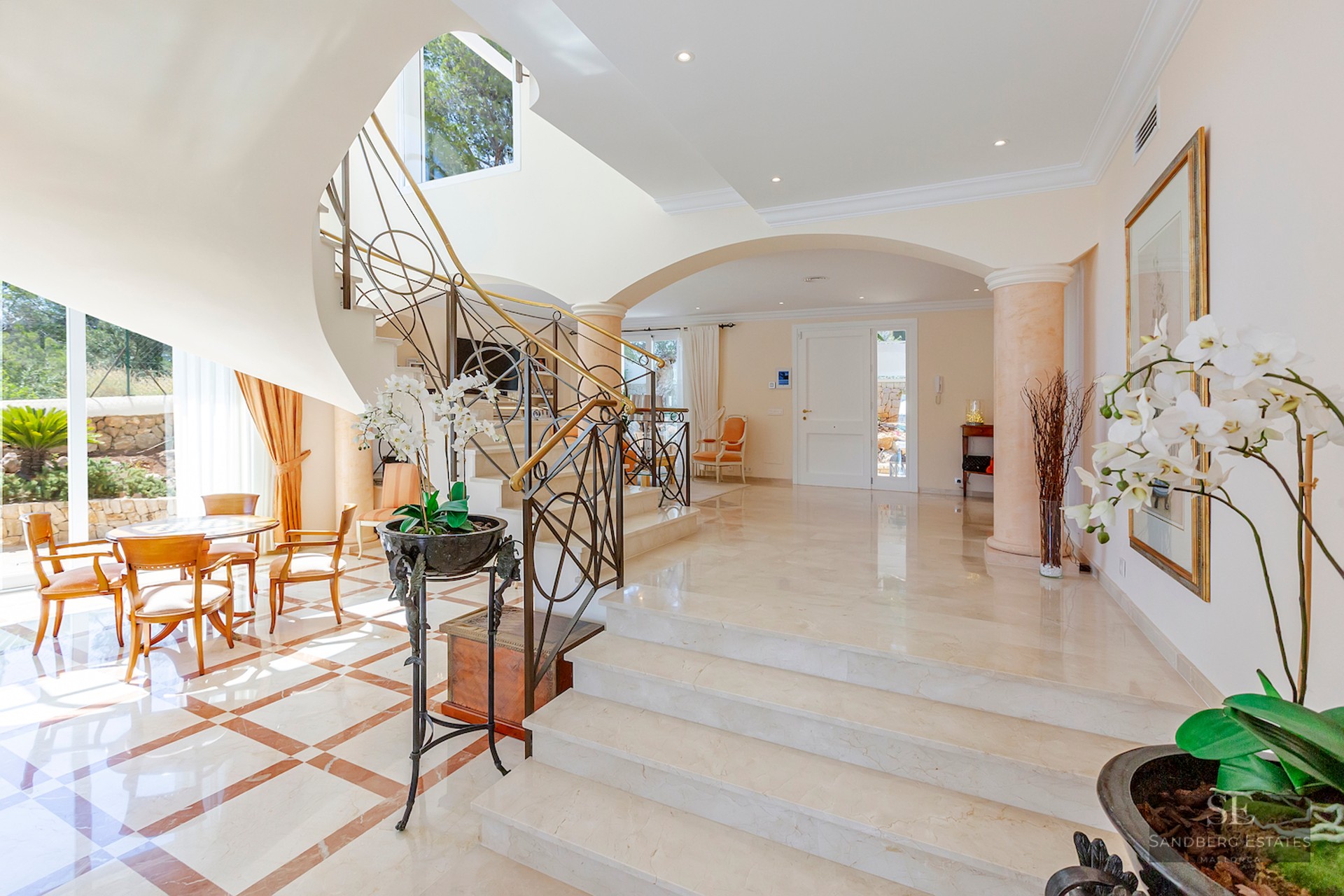 A bright luxury entrance hall featuring a white marble staircase with ornate iron railings and arched architecture.