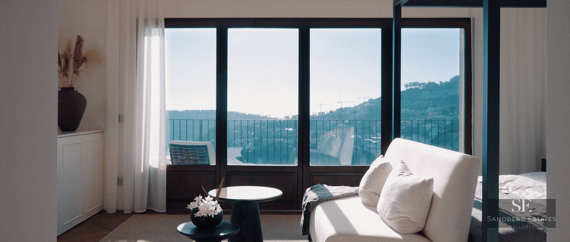 Modern master bedroom featuring a white sofa, canopy bed, and floor-to-ceiling windows overlooking the sea and hills.