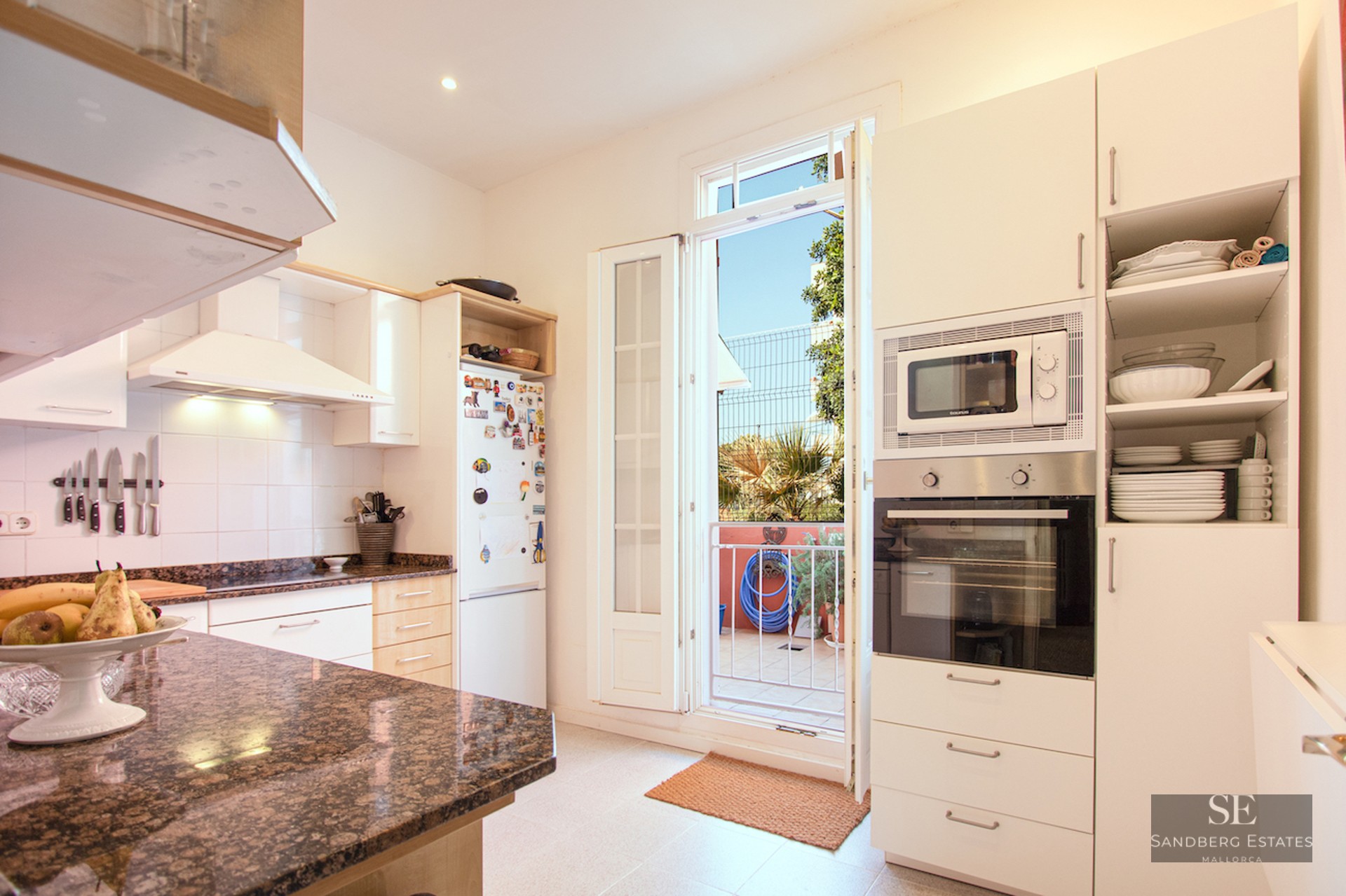 Modern white kitchen featuring brown granite countertops and an open door leading to a sunny balcony.