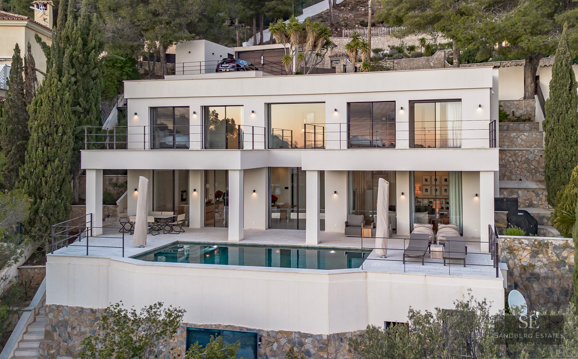 Modern white two-story villa on a hillside with a rectangular pool and multiple terraces at dusk.