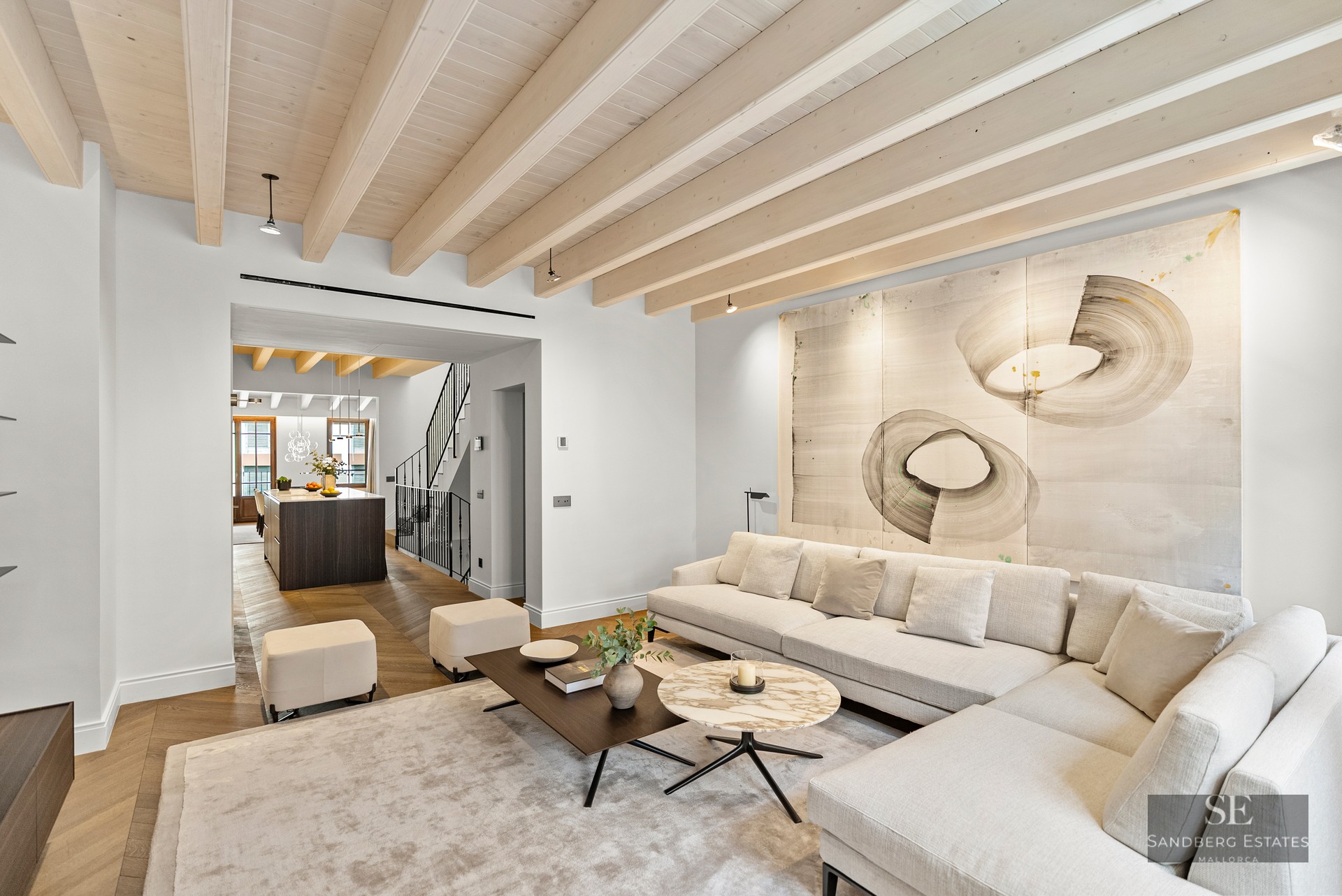 A bright living room with a large white sectional sofa, wooden ceiling beams, and abstract art above the couch.