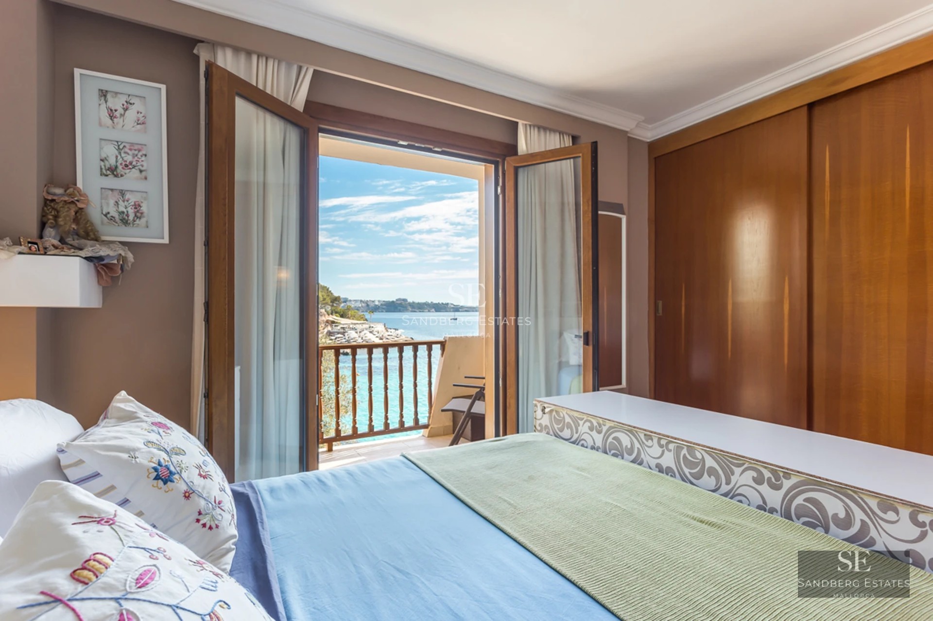 A bedroom with a large bed and open wooden French doors leading to a balcony overlooking the bright blue sea.