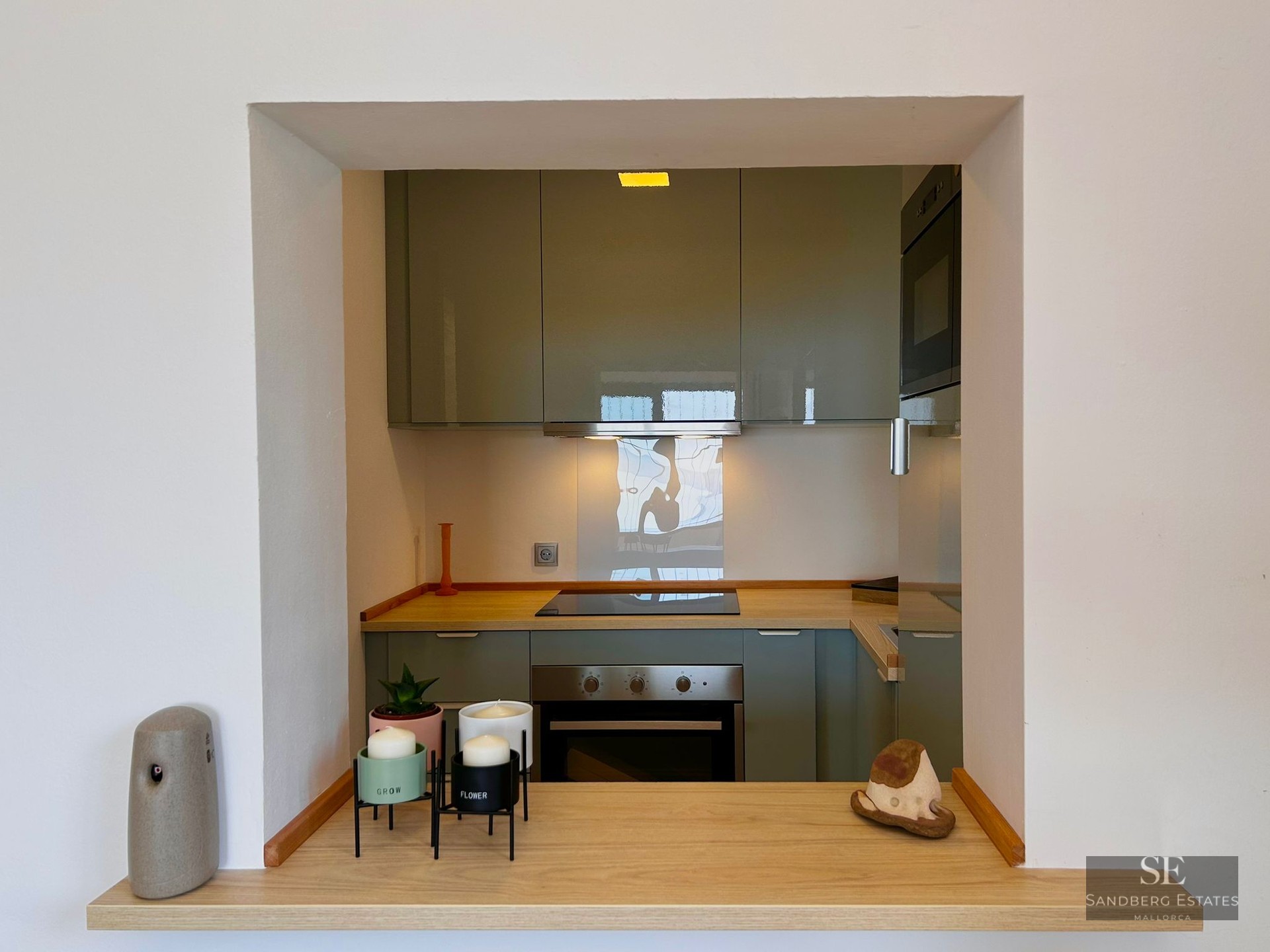 View through a wall opening of a modern kitchen with sage green cabinets and wood countertops.