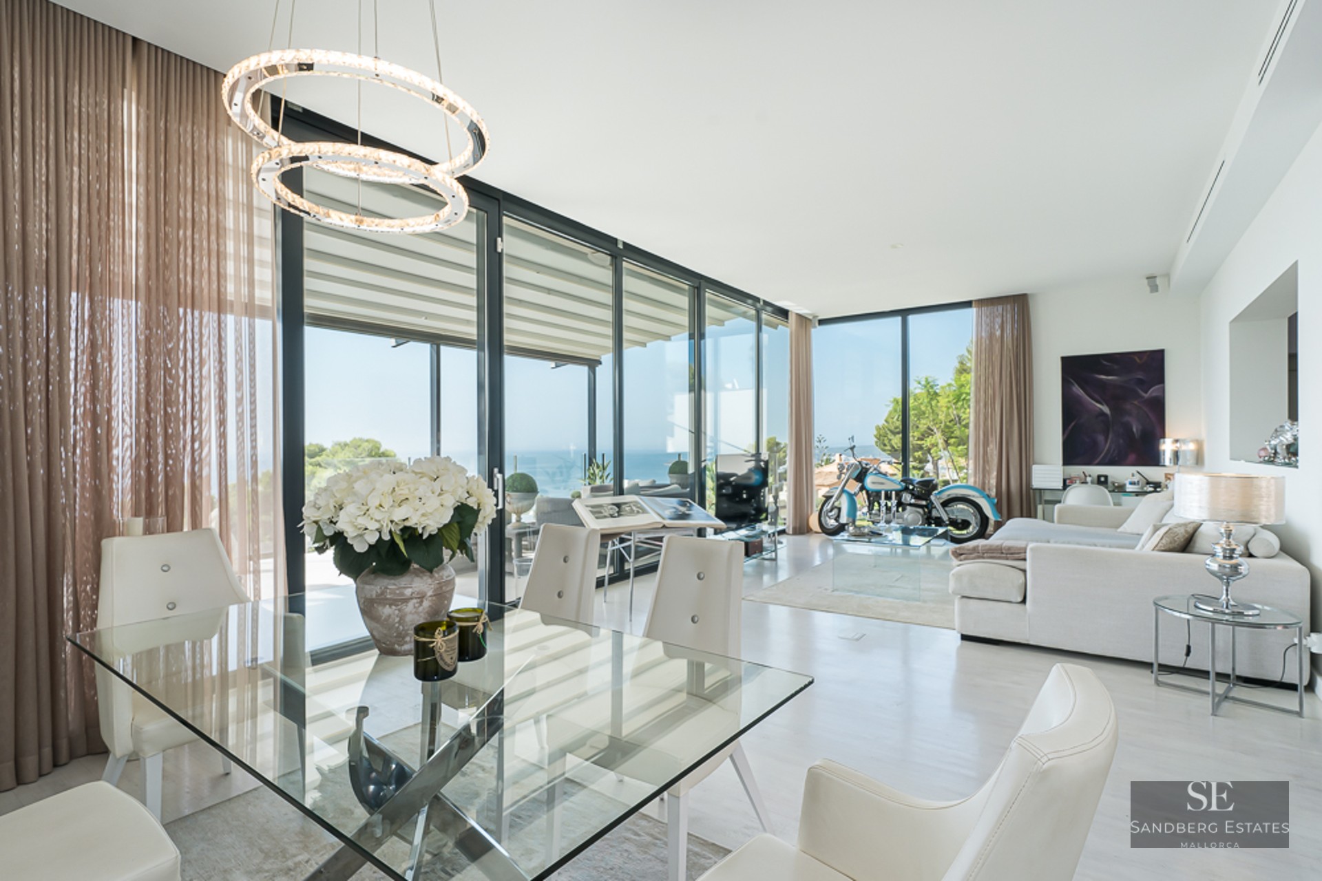 Bright living room with glass dining table, white sofa, floor-to-ceiling windows, and a vintage motorcycle as decor.