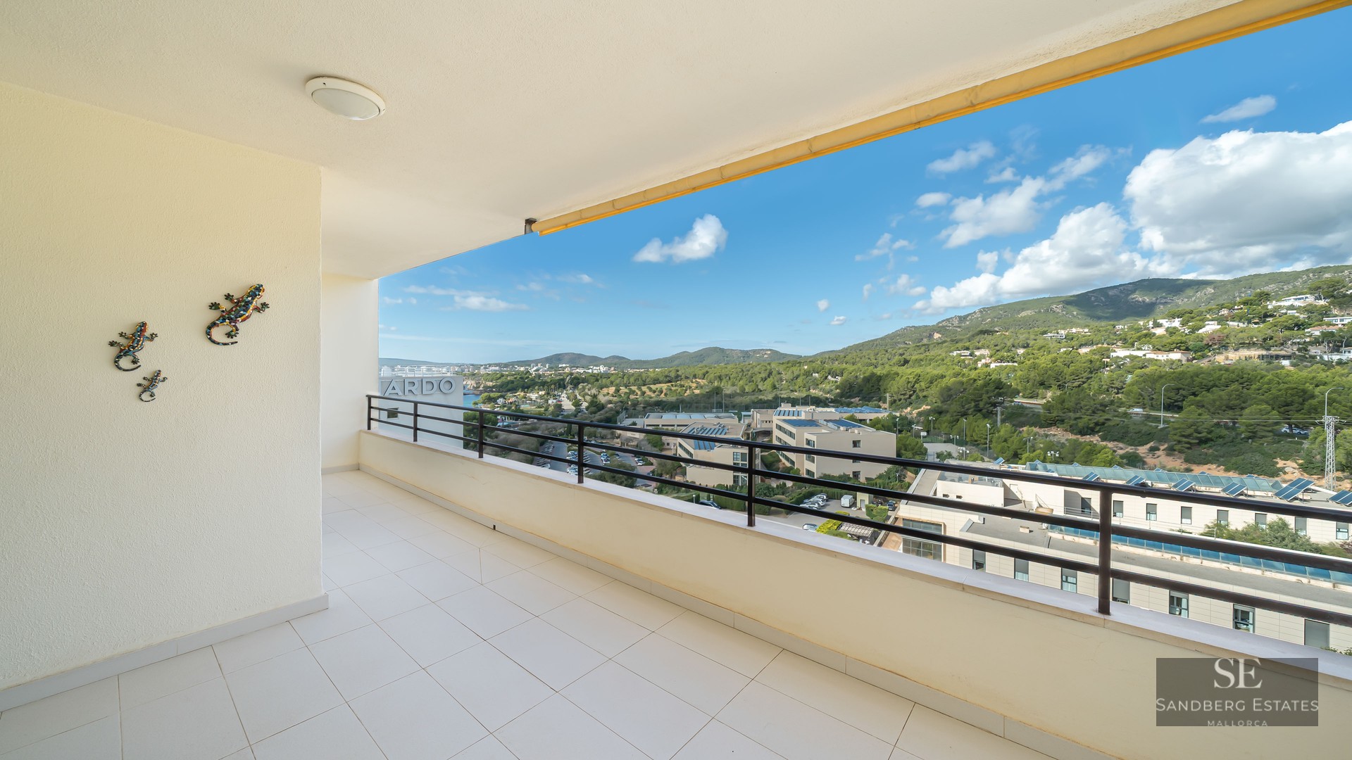 Spacious white tiled balcony with a black metal railing overlooking green hills and a blue sky with white clouds.