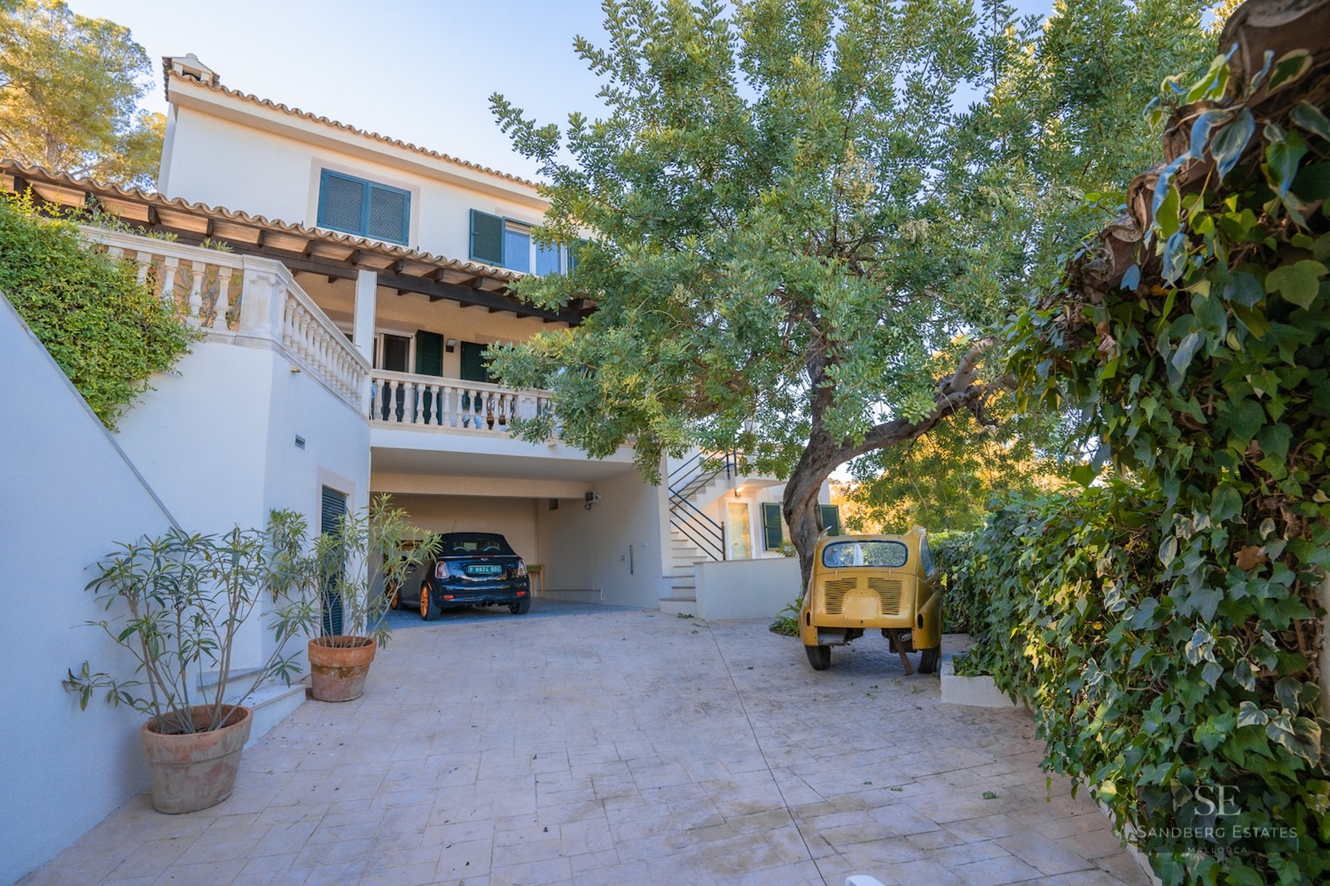 Paved driveway leading to a covered parking space at a white villa with green shutters and mature trees.