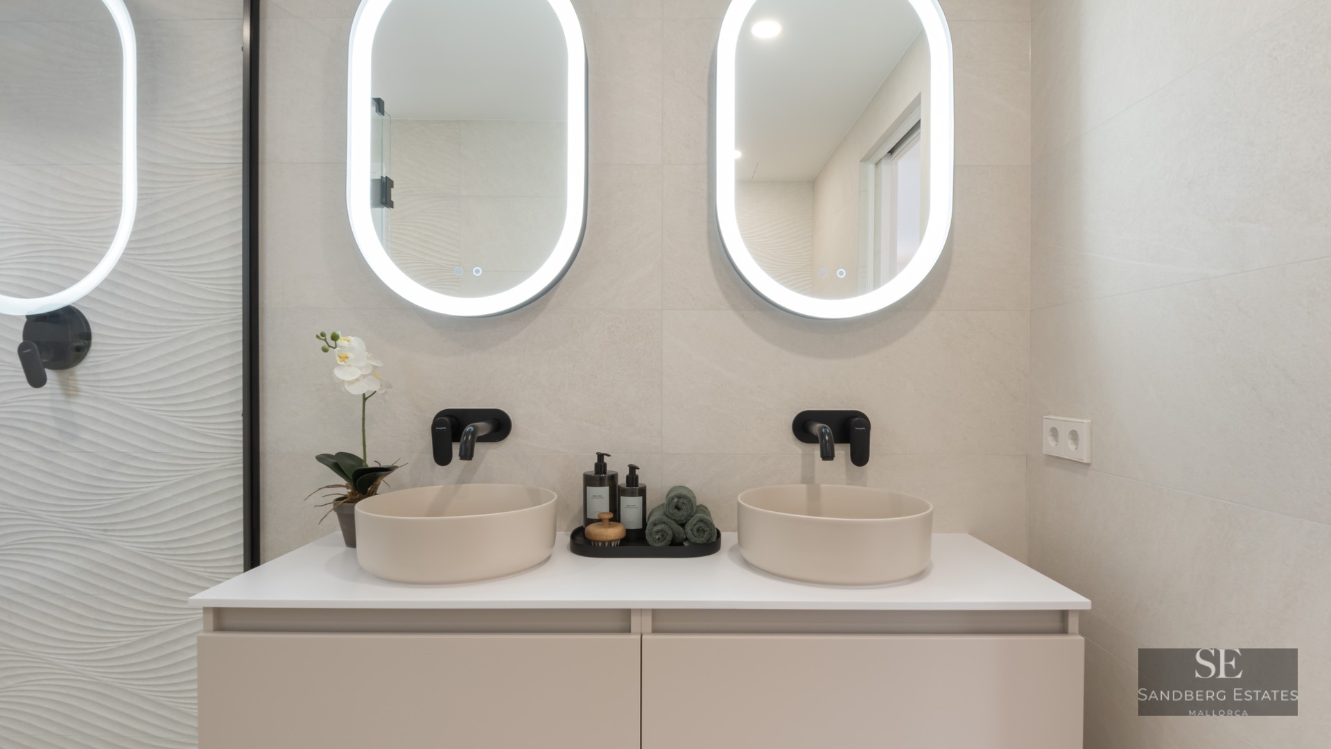Modern bathroom with two round vessel sinks, black faucets, and oval backlit mirrors on a beige wall.