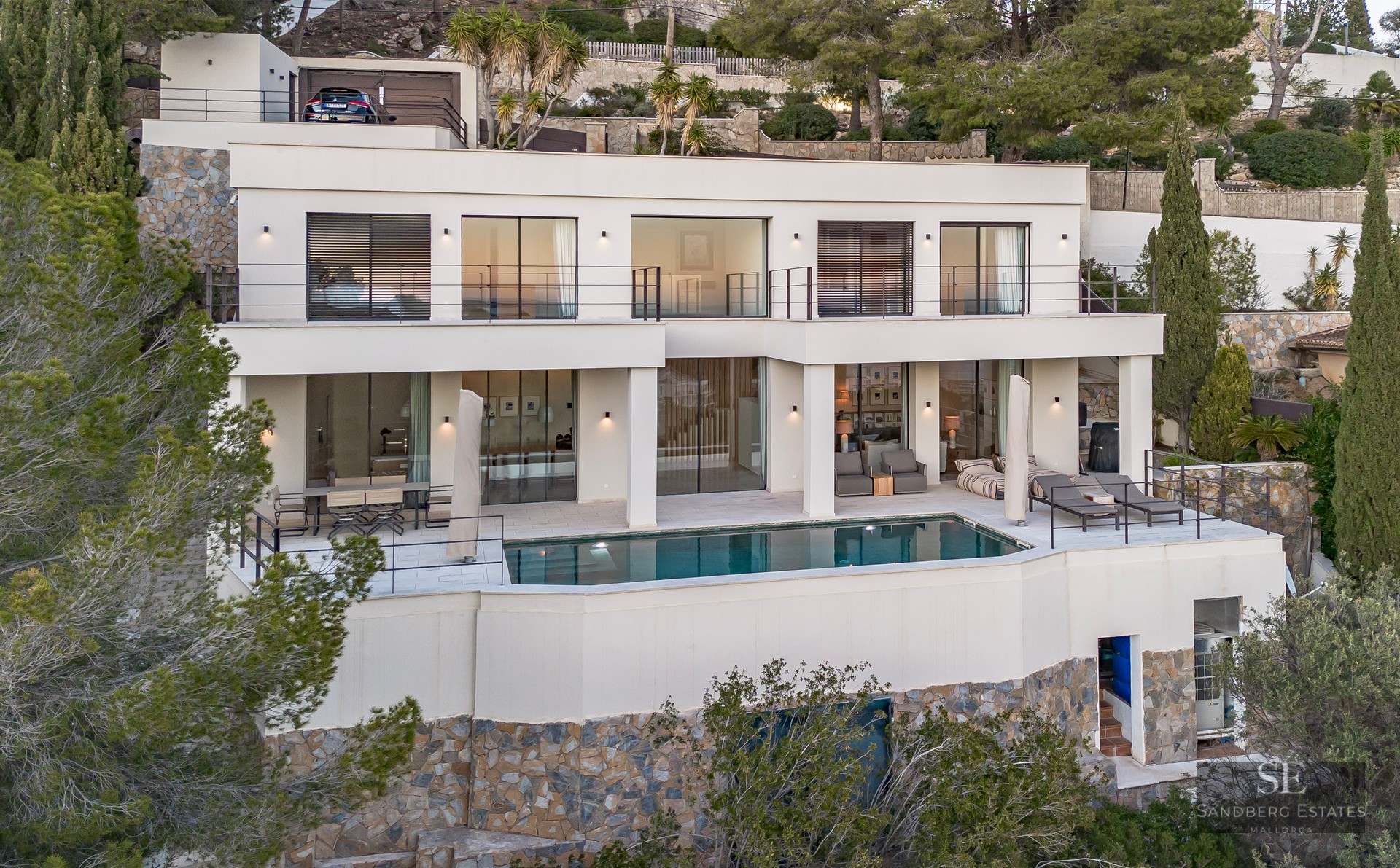 Two-story white modern villa with an infinity pool, spacious terrace, and lush green surroundings on a hillside.