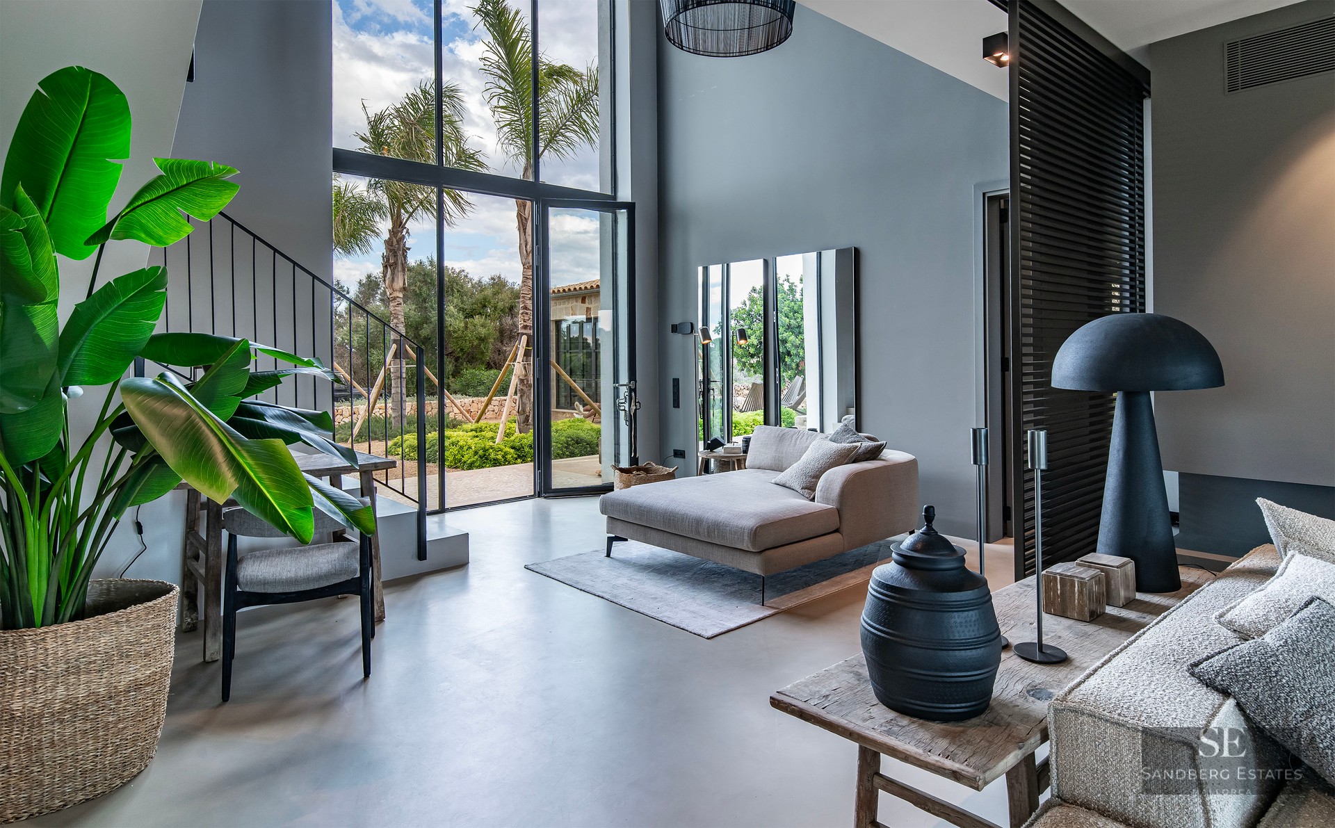 Spacious living area with high ceilings, floor-to-ceiling windows with palm tree views, and modern grey chaise lounge.