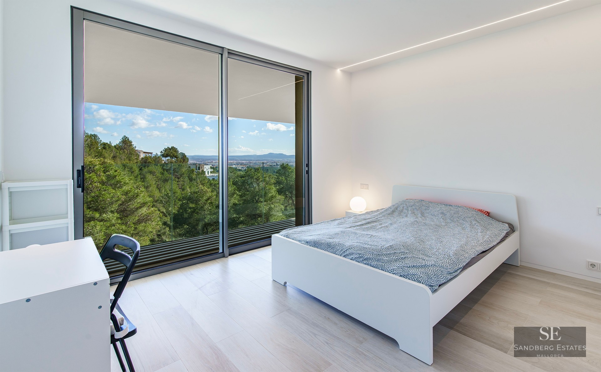 Bright bedroom featuring a white bed, light wood flooring, and large glass doors overlooking a forest and mountains.