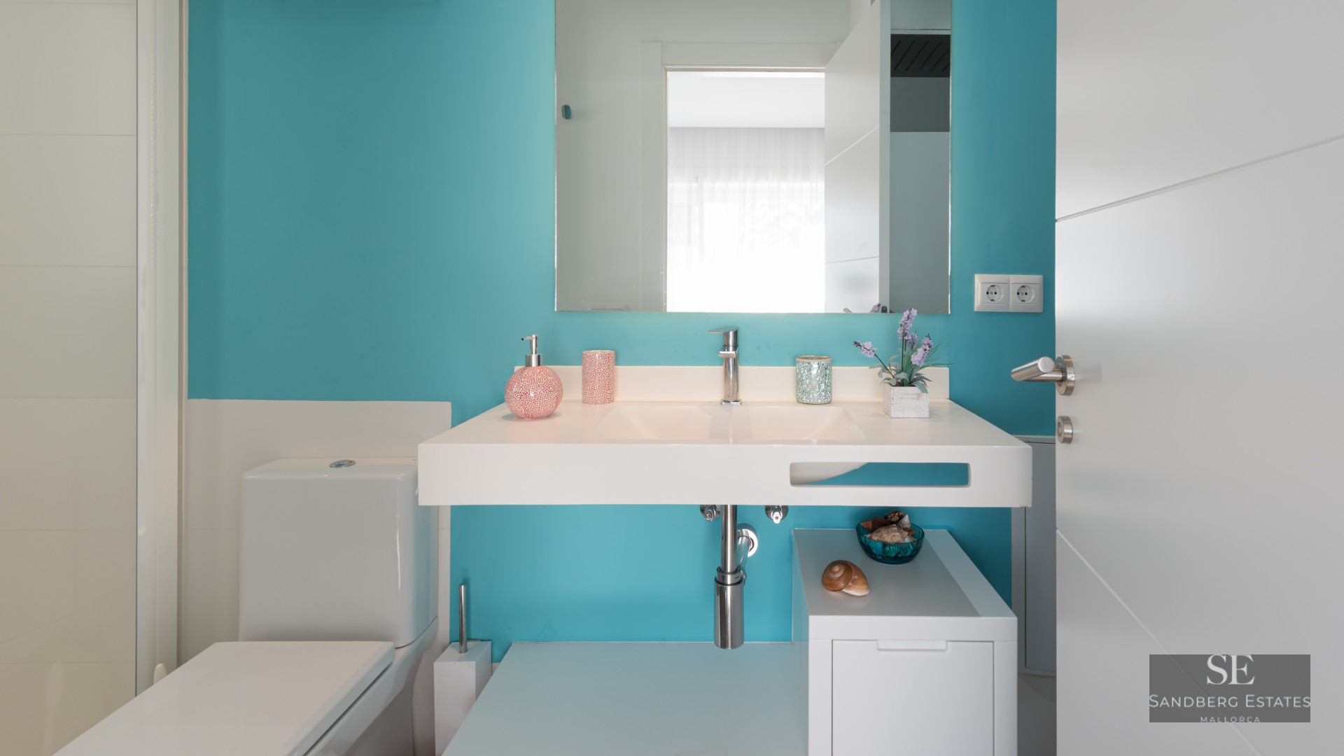 Salle de bain moderne avec mur turquoise, vasque suspendue blanche, grand miroir et toilettes blanches.
