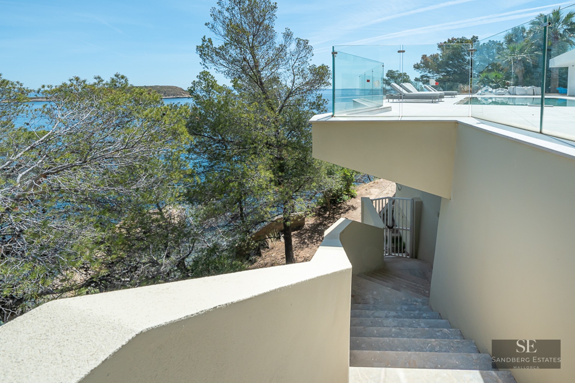 Luxury pool terrace with glass railings and stone stairs overlooking the turquoise Mediterranean Sea and pine trees.