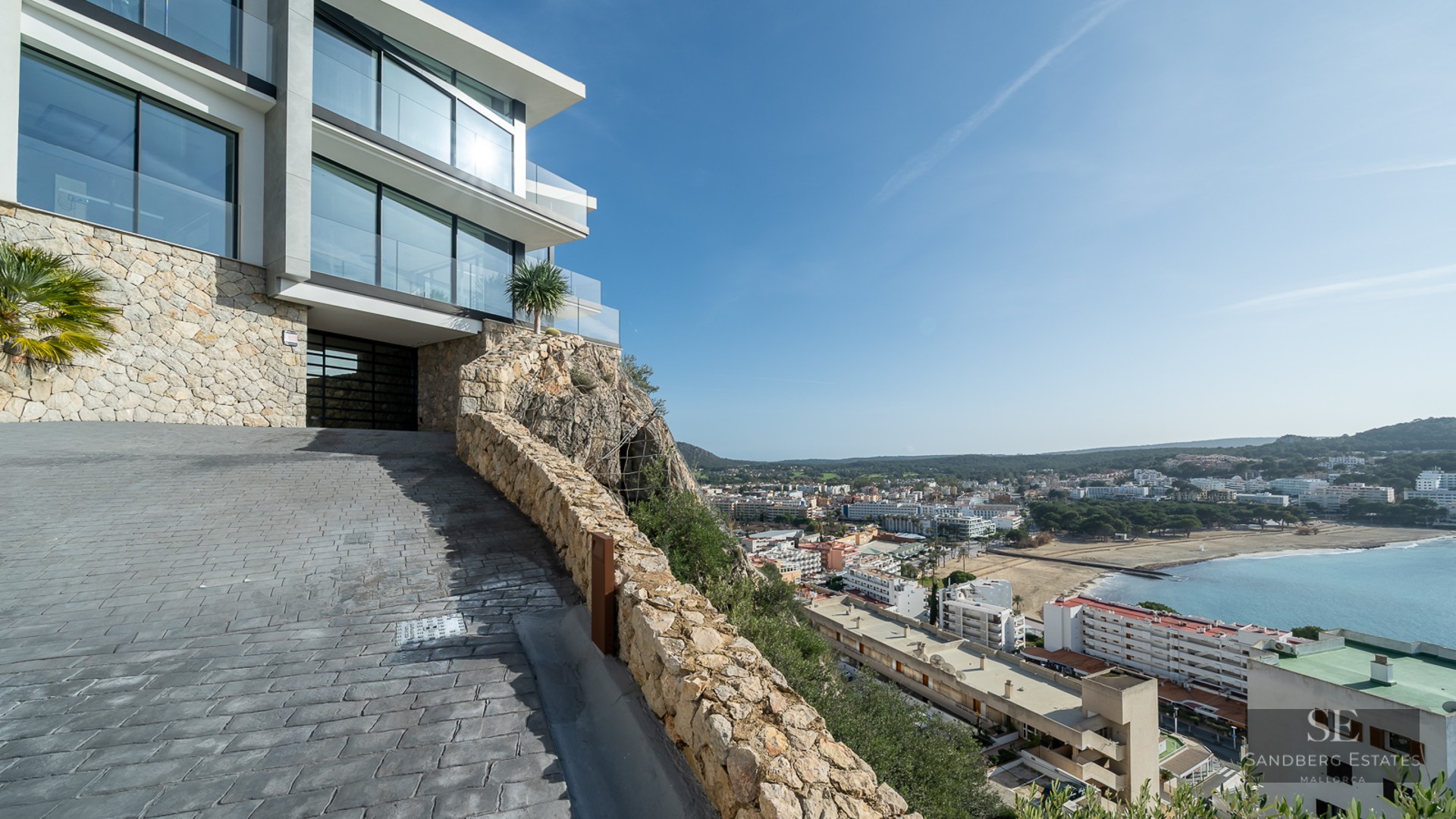 Modern villa with glass walls and stone facade on a cliff overlooking a coastal bay and town under a clear blue sky.