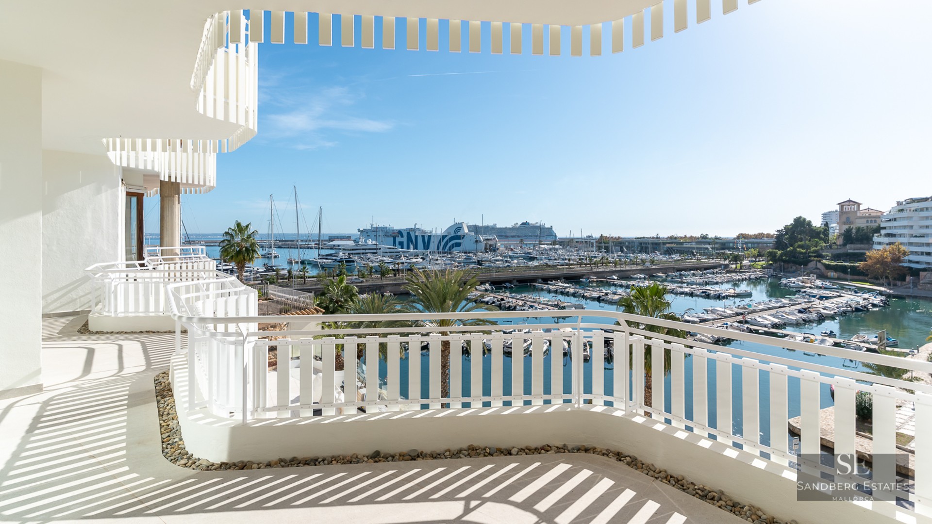 White curved balcony with modern railing overlooking a sunny marina with yachts and palm trees.