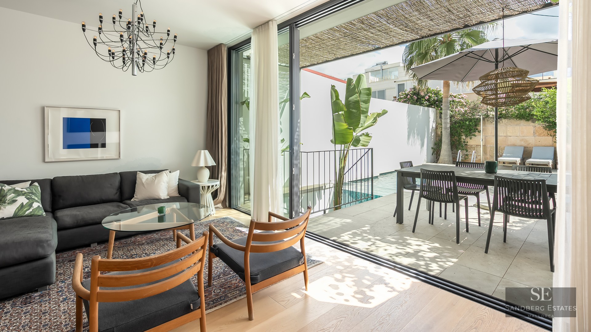 Bright living room with dark sofa and wooden chairs opening to a sunny terrace with a swimming pool and dining area.