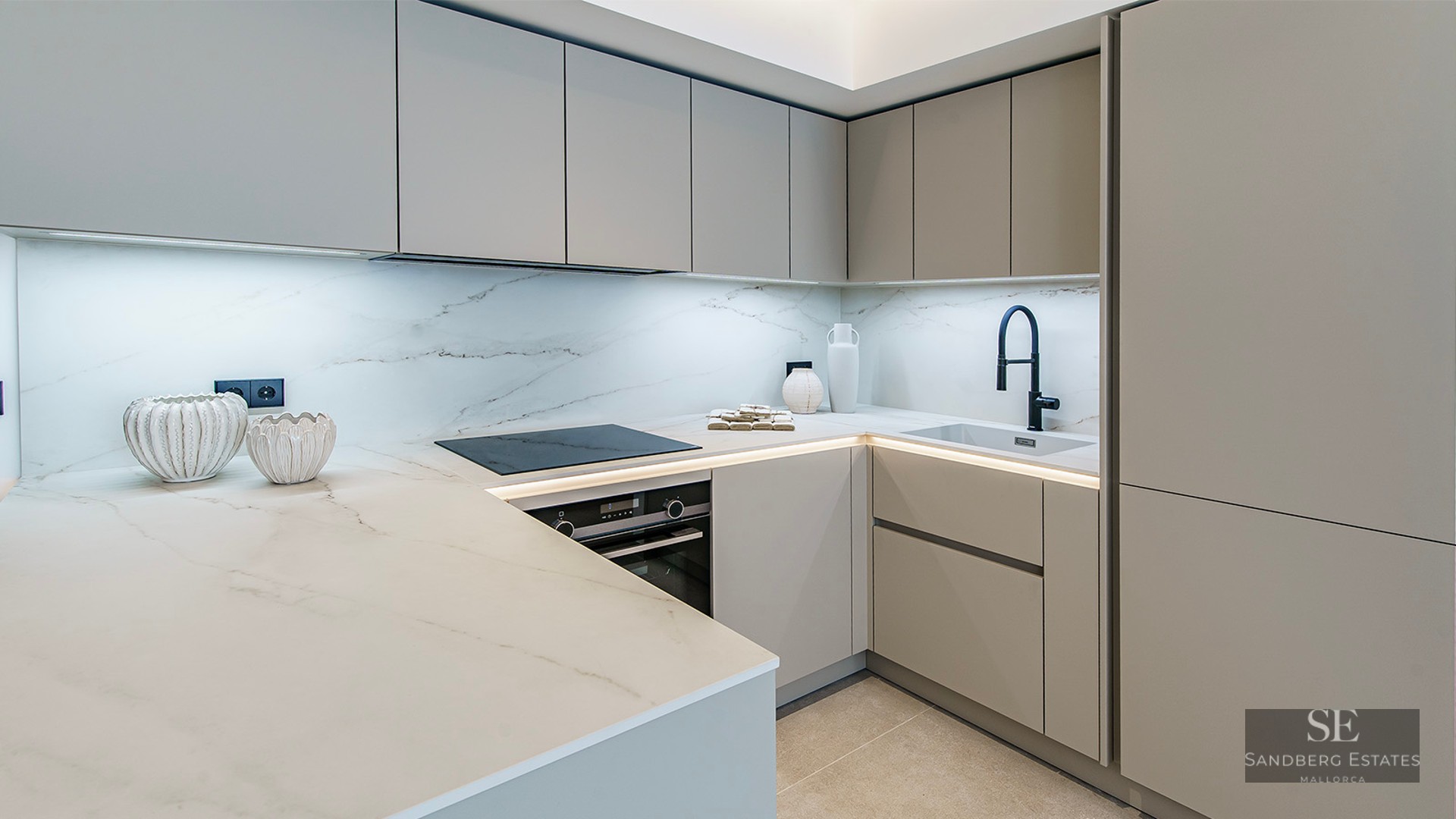A modern kitchen featuring light gray cabinetry, white marble countertops, LED lighting, and a matte black faucet.