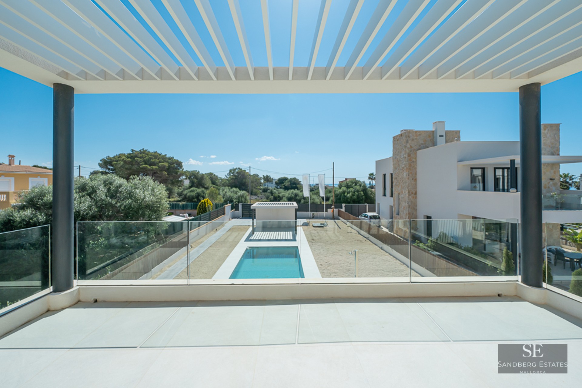 View from a modern balcony with a white pergola and glass railing overlooking a rectangular swimming pool and garden.