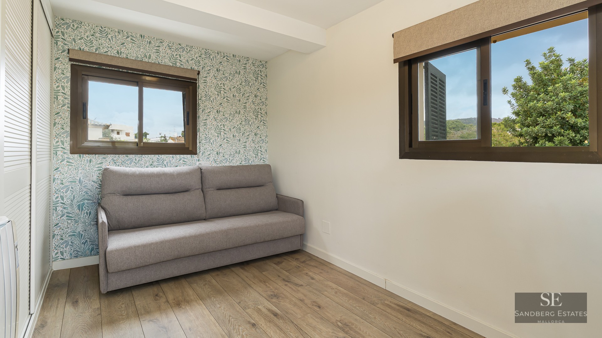 Modern bedroom featuring a grey sofa, botanical leaf pattern wallpaper, wooden floors, and natural light from two windows.
