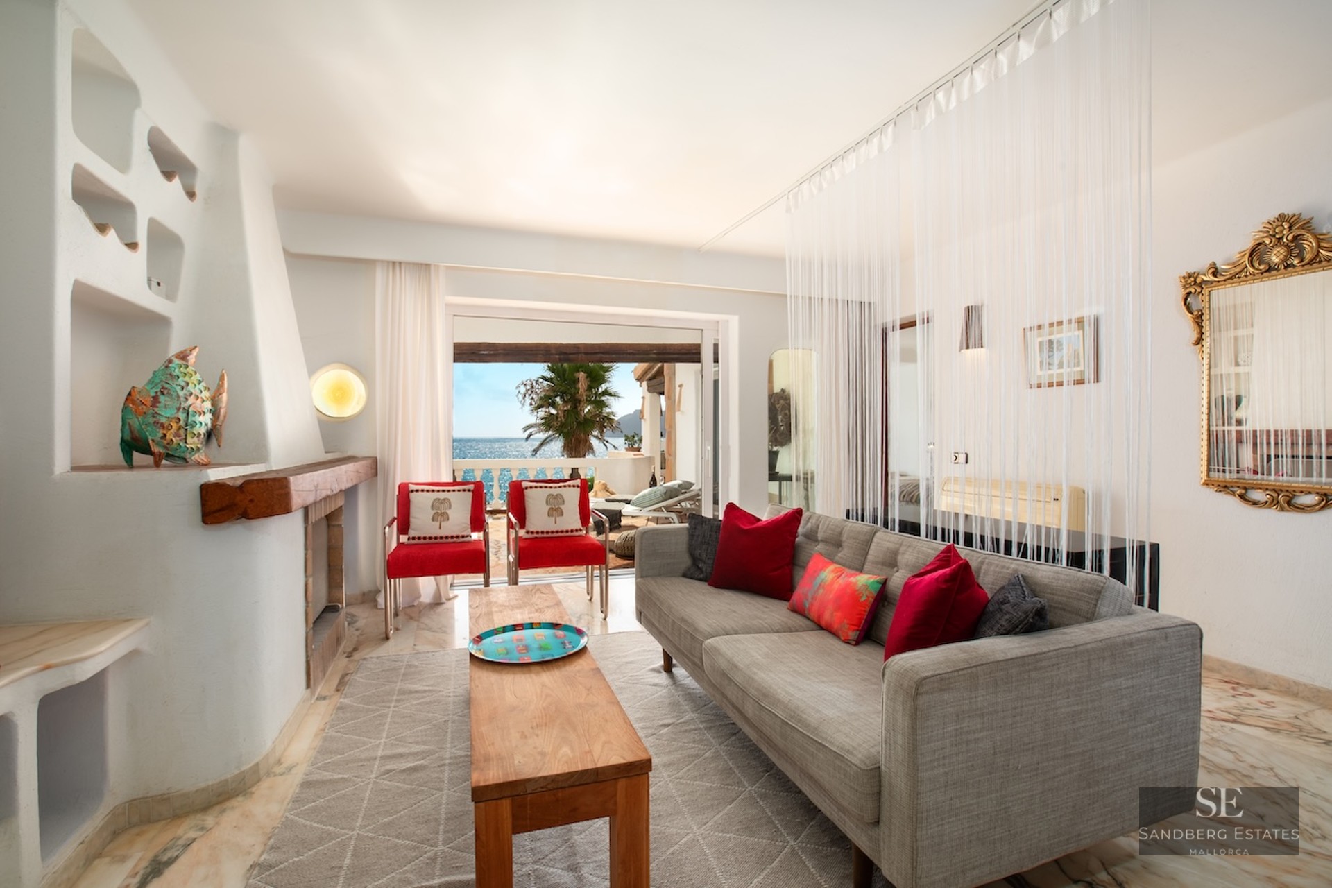 Living room featuring a grey sofa, red chairs, rustic fireplace, and large windows overlooking the sea and terrace.