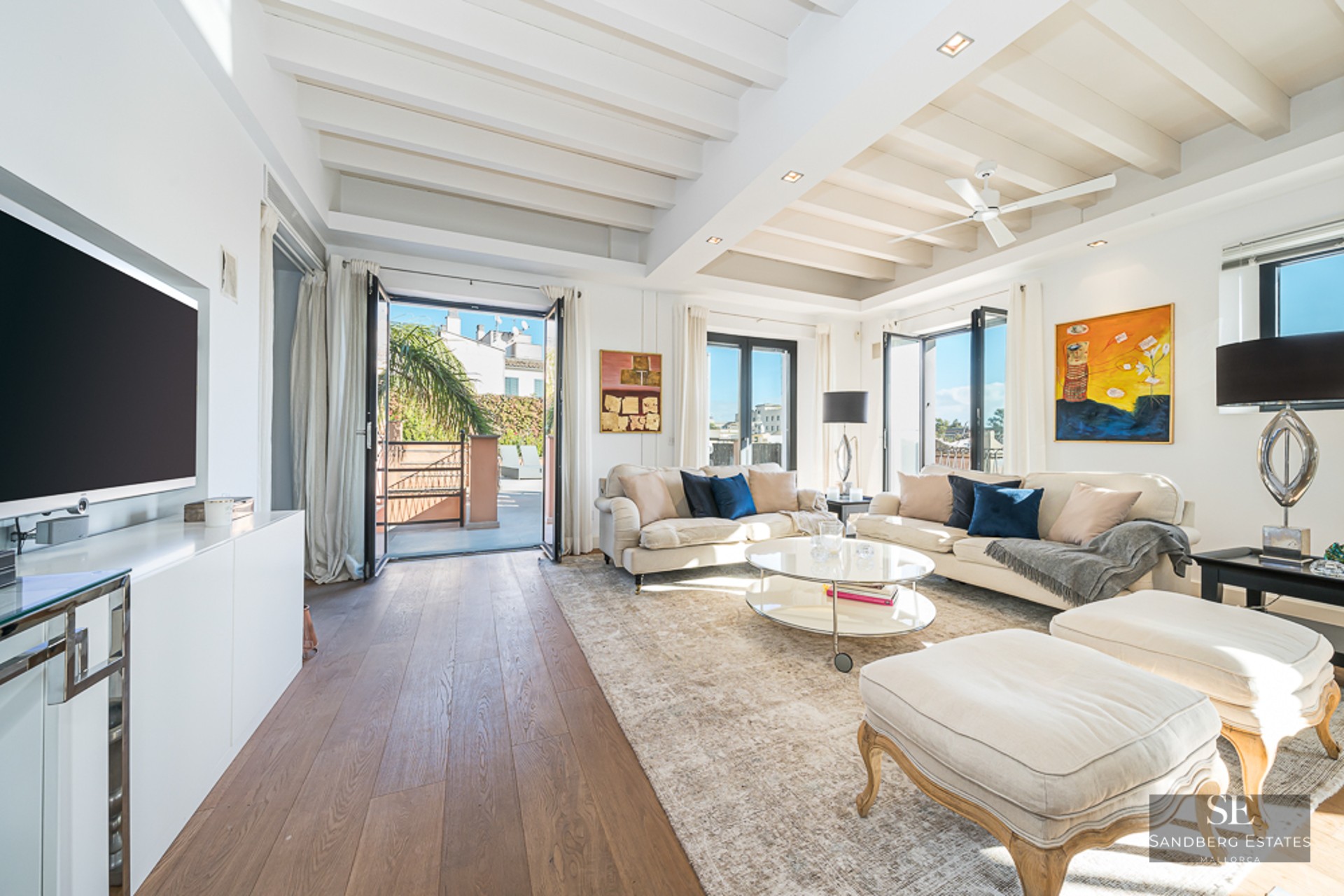 Elegant living room featuring cream sofas, white beamed ceilings, and access to a sunny outdoor terrace.