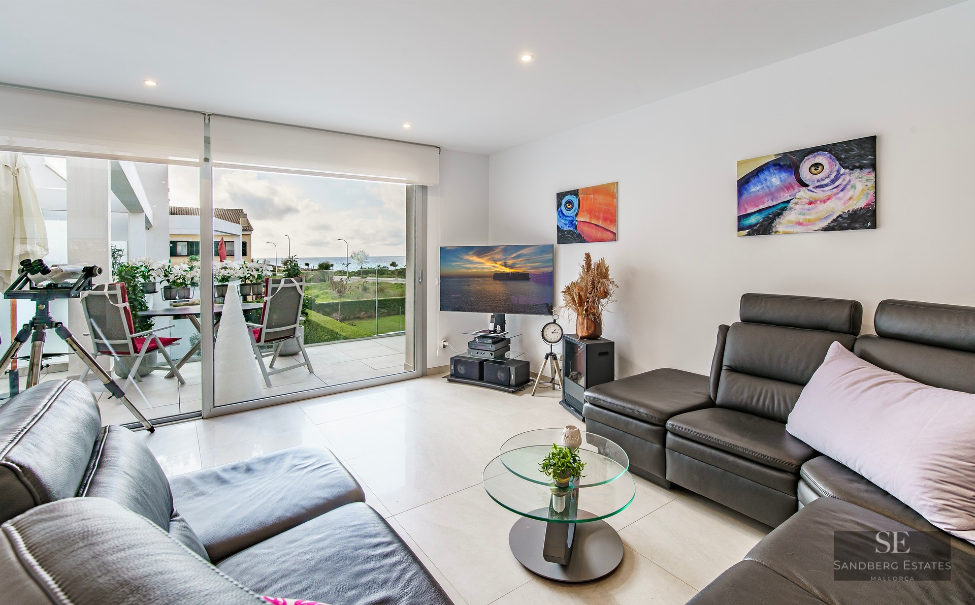 Modern living room with black leather sofas, glass coffee table, and large sliding doors opening to a terrace with sea views.