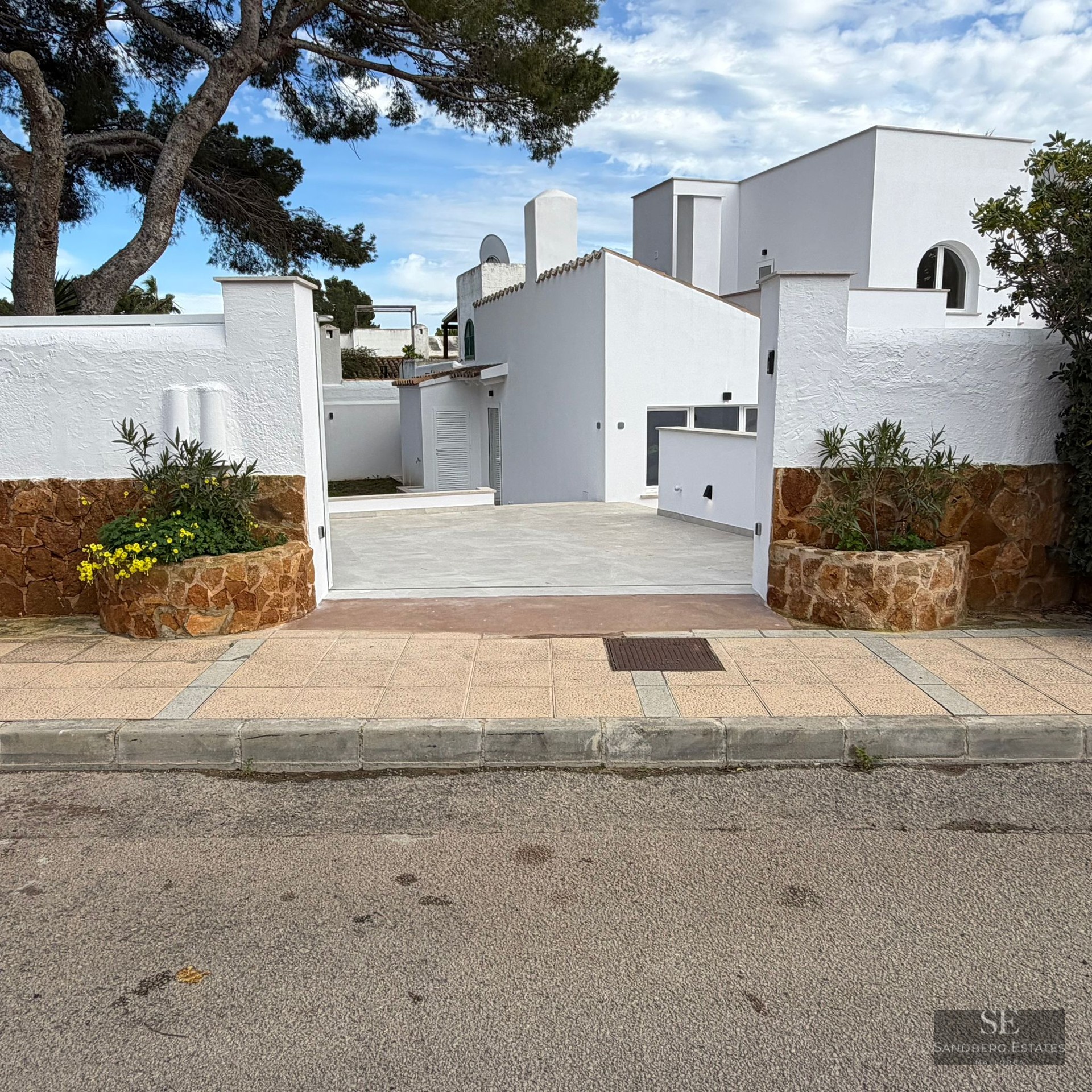 Entrance to a white villa featuring stone-clad walls, planters with yellow flowers, and a paved driveway under pine trees.