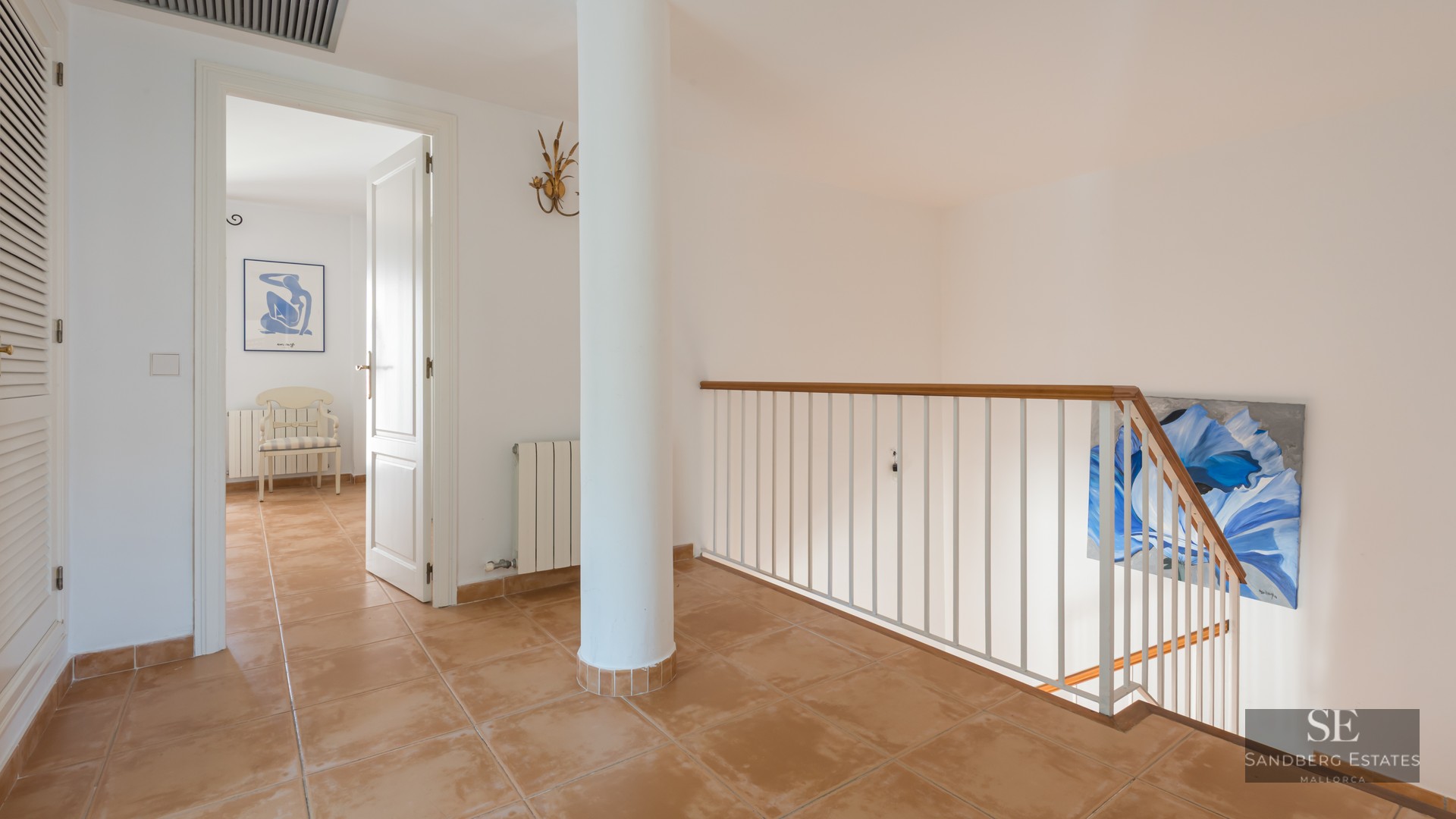 Hallway with terracotta tiles, a white pillar, and a staircase railing, leading to an open doorway.