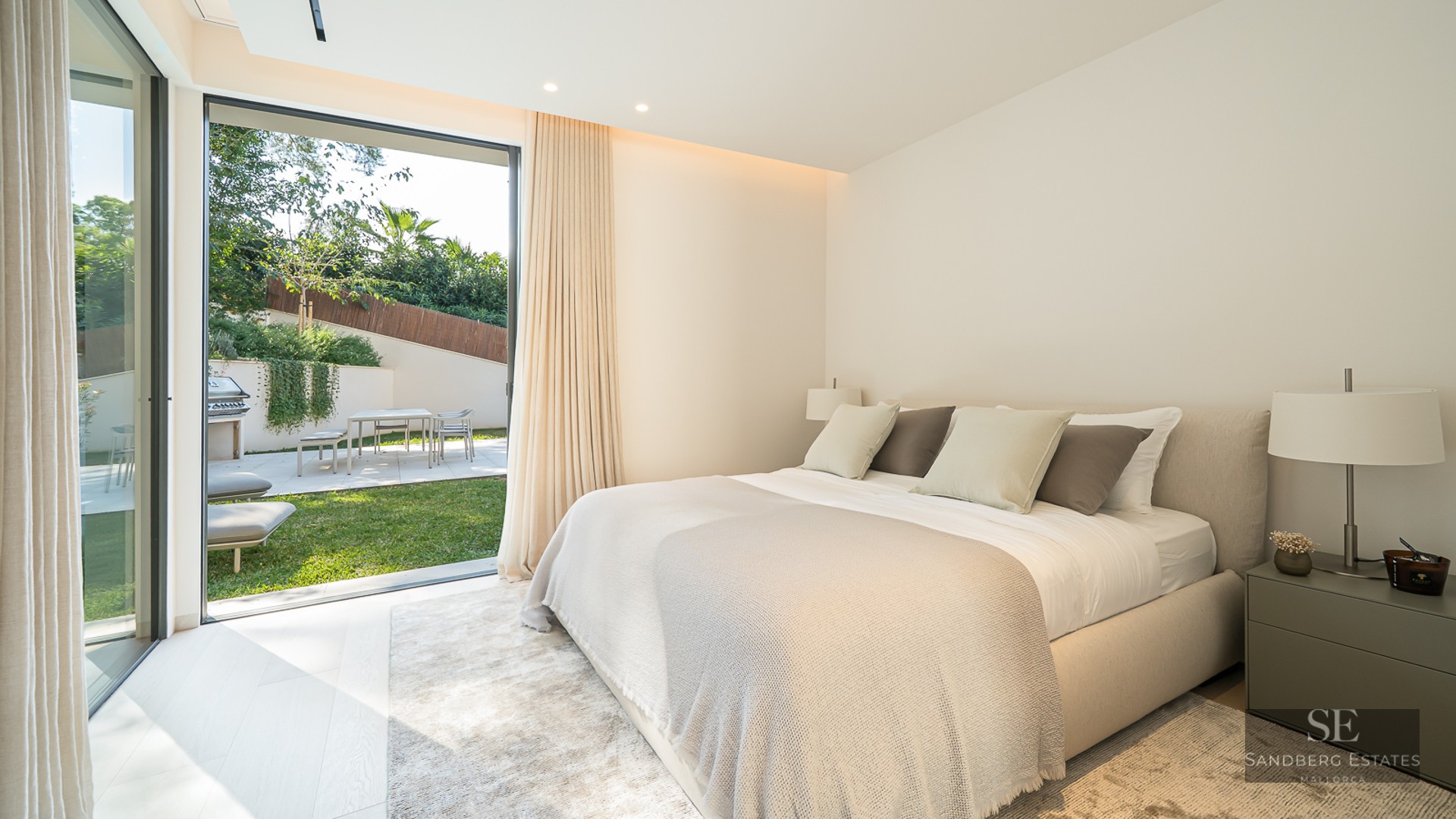 A bright, modern bedroom with a large bed opening onto a sunny private garden through glass sliding doors.