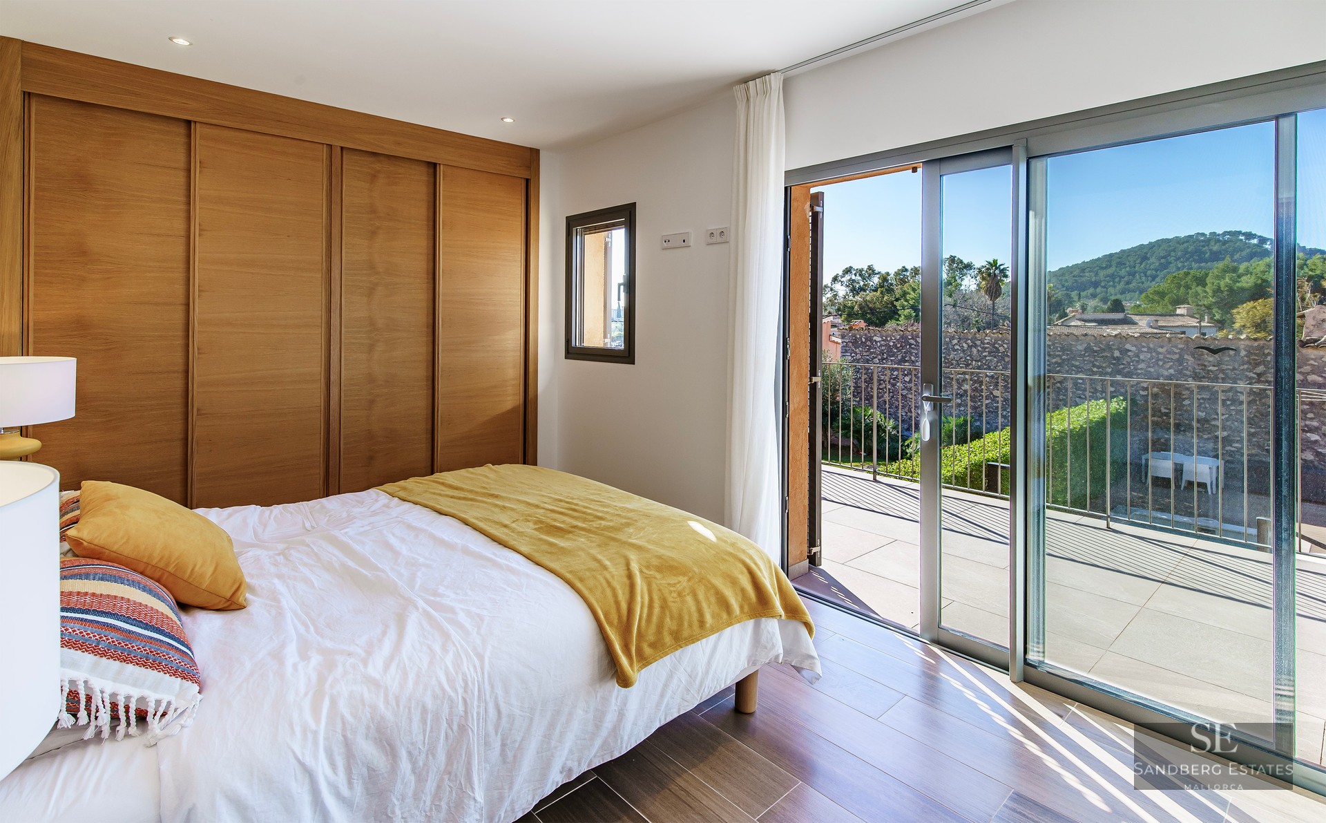 Bright bedroom with wooden built-in closets and large sliding glass doors opening onto a sunny terrace with green views.